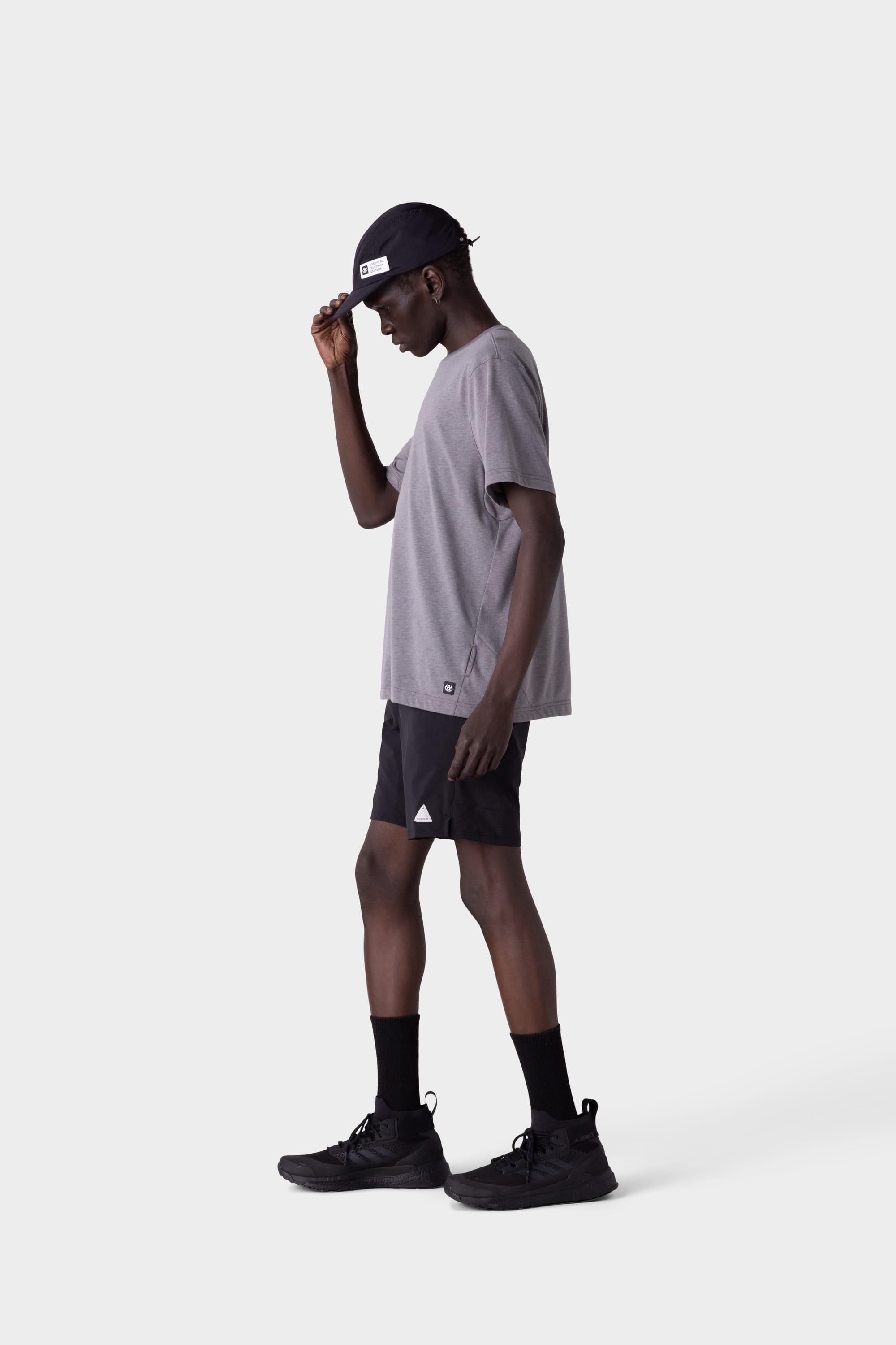 A person in a gray T-shirt, 686 Mens ATP Stretch Performance Short by 686, black socks, and black sneakers stands in profile against a plain light background, adjusting a black cap with one hand.