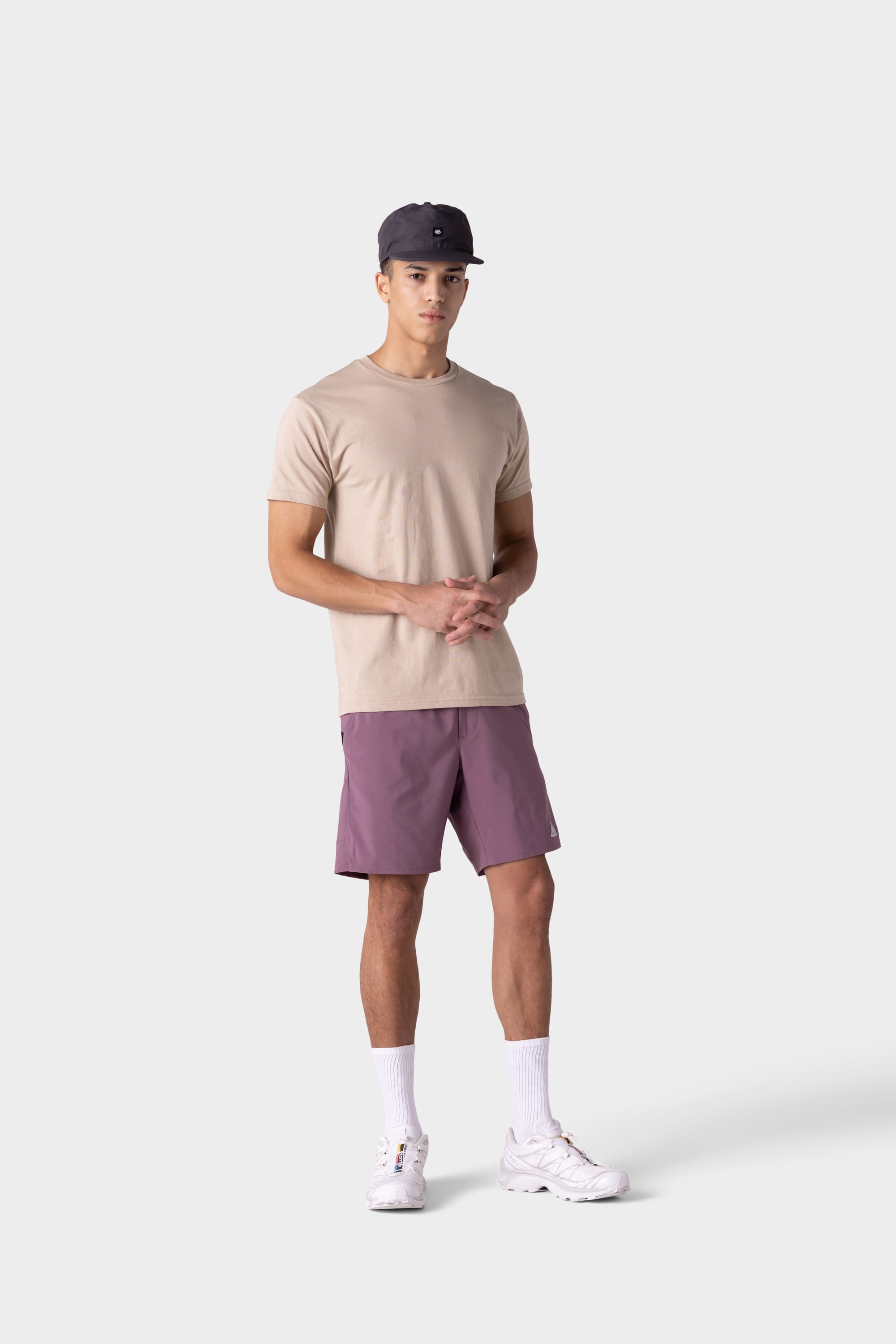 A young man stands against a plain background, wearing beige T-shirt, white socks and sneakers, a dark baseball cap, and 686 Mens ATP Stretch Performance Shorts by 686. His hands are clasped in front of him as he looks forward.