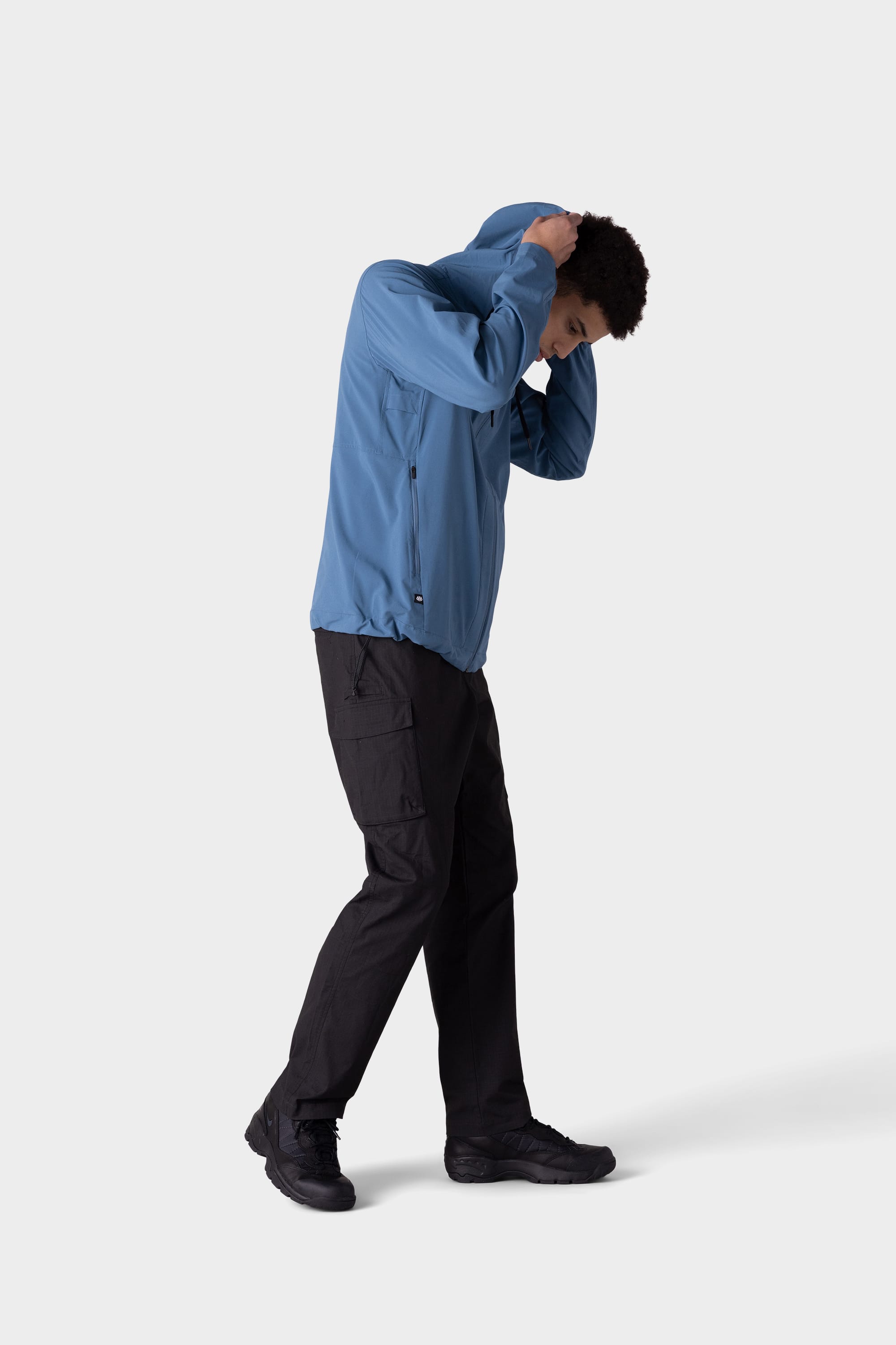 A person in a 686 Mens ATP Stretch Wind Jacket pulls the hood over their head while looking down, paired with black cargo pants and shoes, standing against a plain light background.