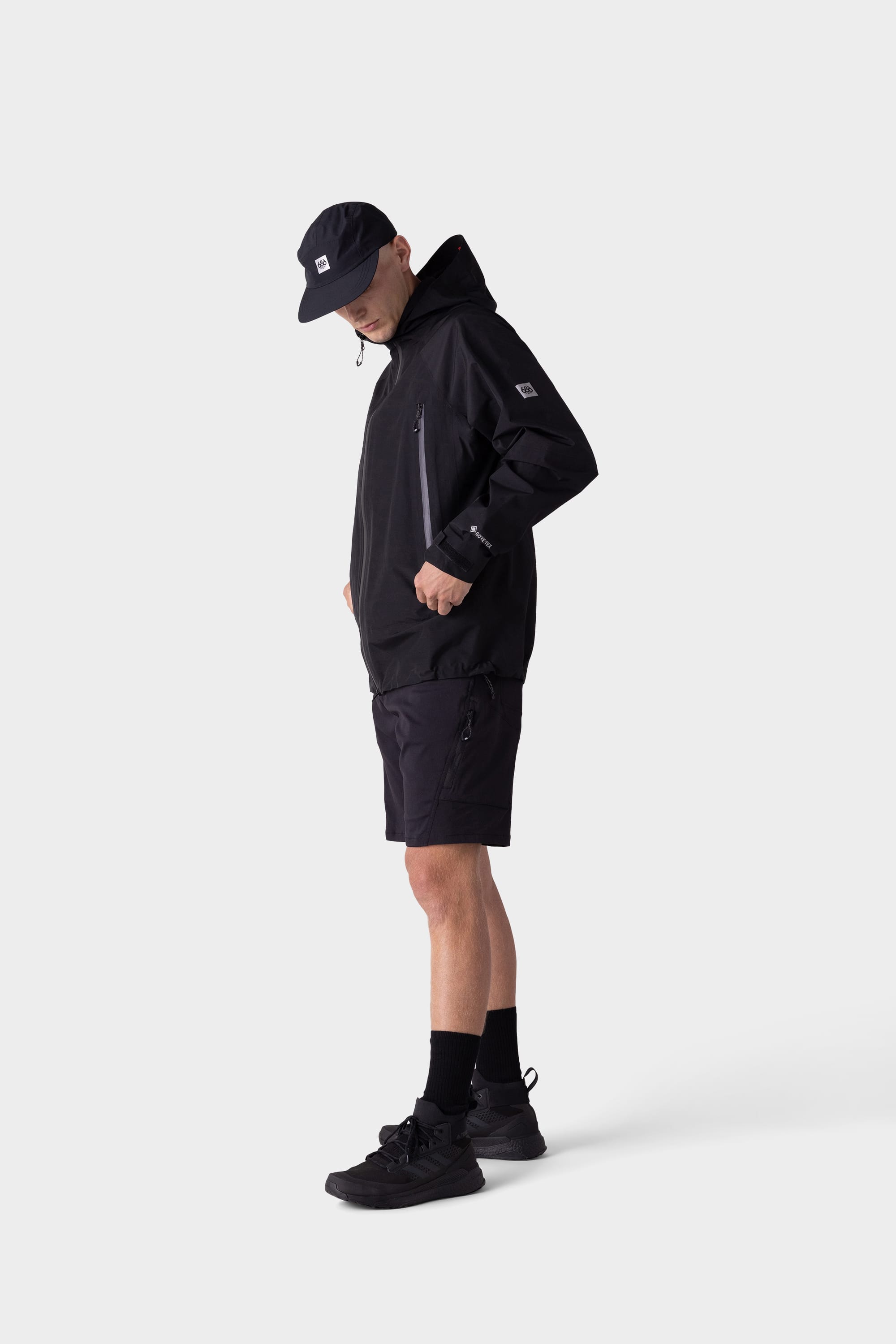 A person in a black hooded jacket, 686 Mens Anything Hybrid Cargo Short by 686, black socks, sneakers, and cap stands against a plain white background with hands in jacket pockets—a minimalist statement.