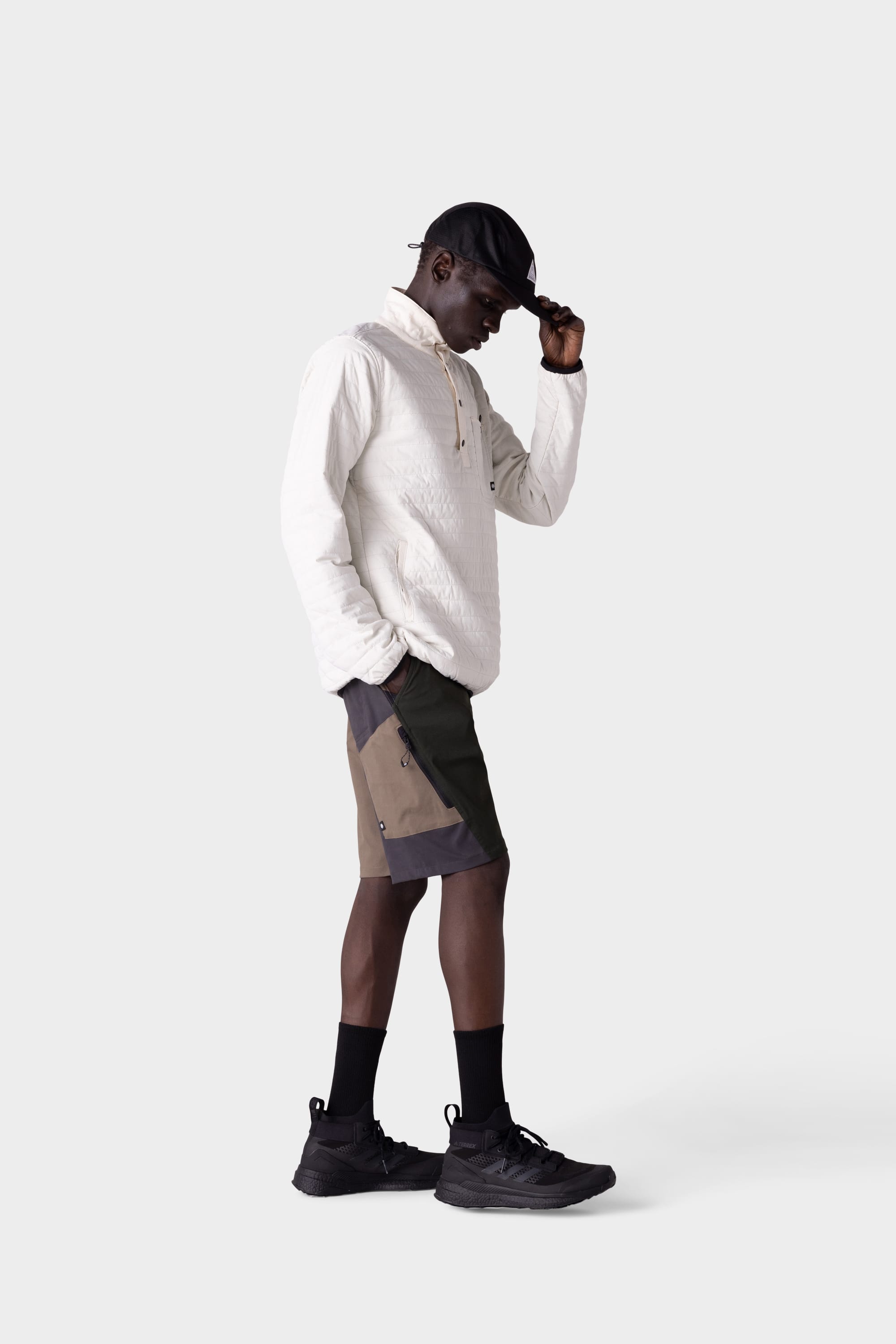 A man stands sideways in a white long-sleeve shirt and 686 Mens Anything Hybrid Cargo Short, wearing black sneakers, socks, and a cap—adjusting the brim while looking down—showcasing minimalist style against a plain light background.