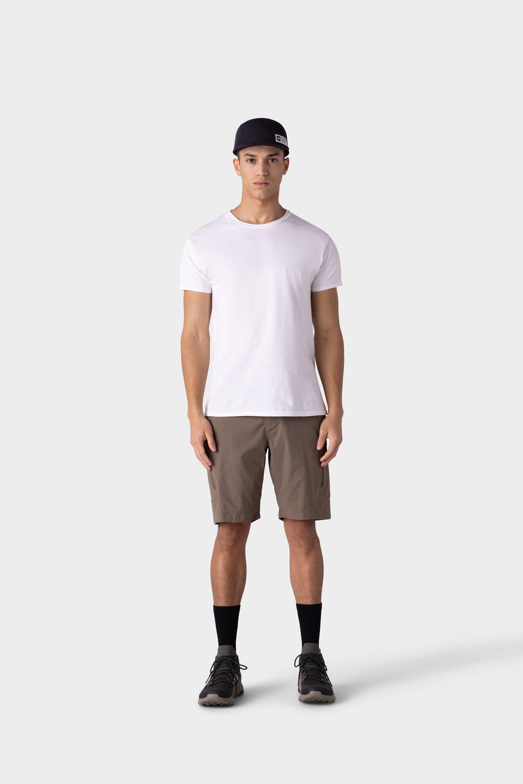 A person in a minimalist design wears a black cap, white t-shirt, 686 Mens Anything Hybrid Cargo Short by 686 in brown, black crew socks, and dark sneakers against a plain gray background.