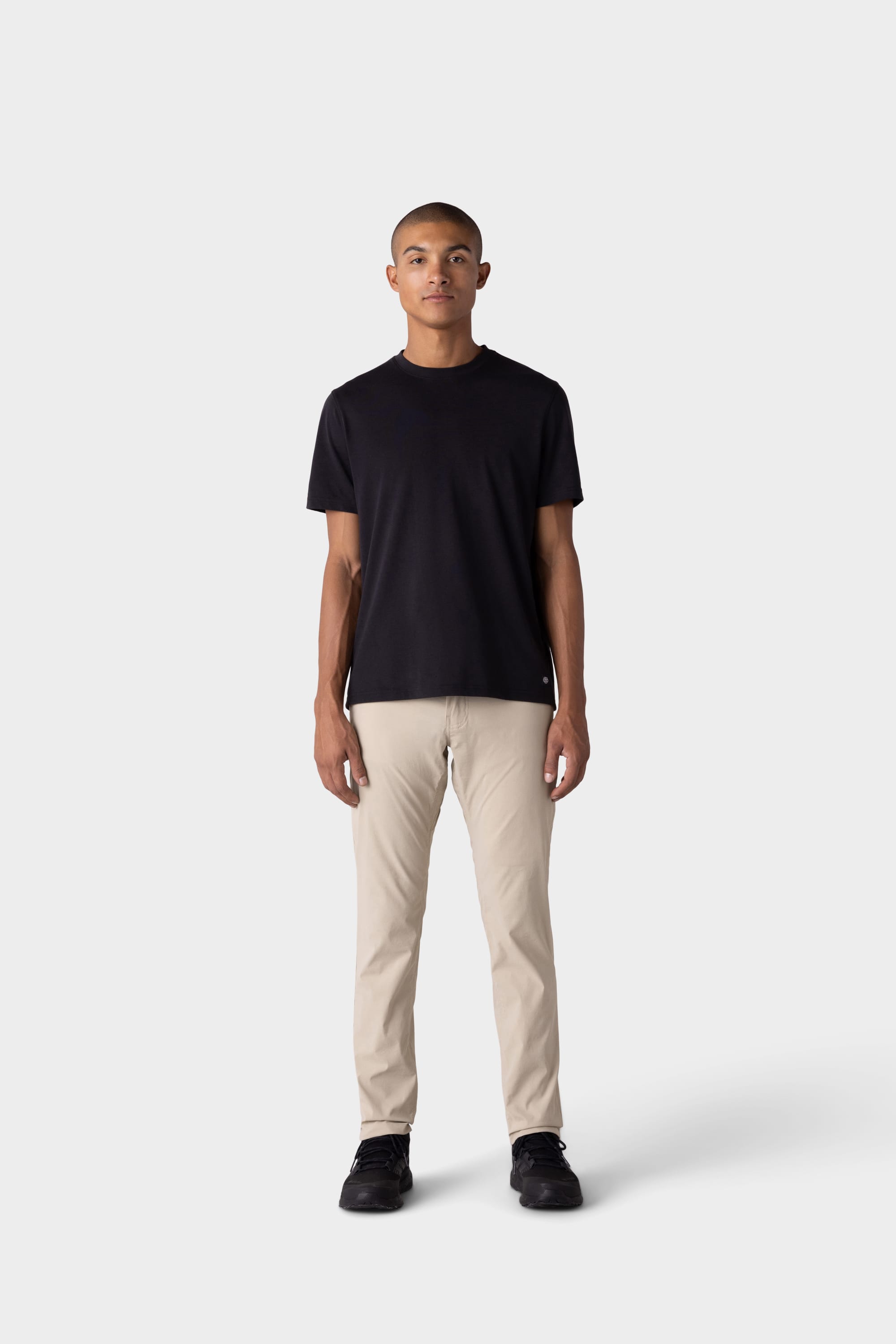 A man stands facing forward against a plain light grey background, wearing a black t-shirt, 686 Mens Everywhere Featherlight Chino Pant - Slim Fit in beige, and black shoes—ready to refresh his summer wardrobe with lightweight style.