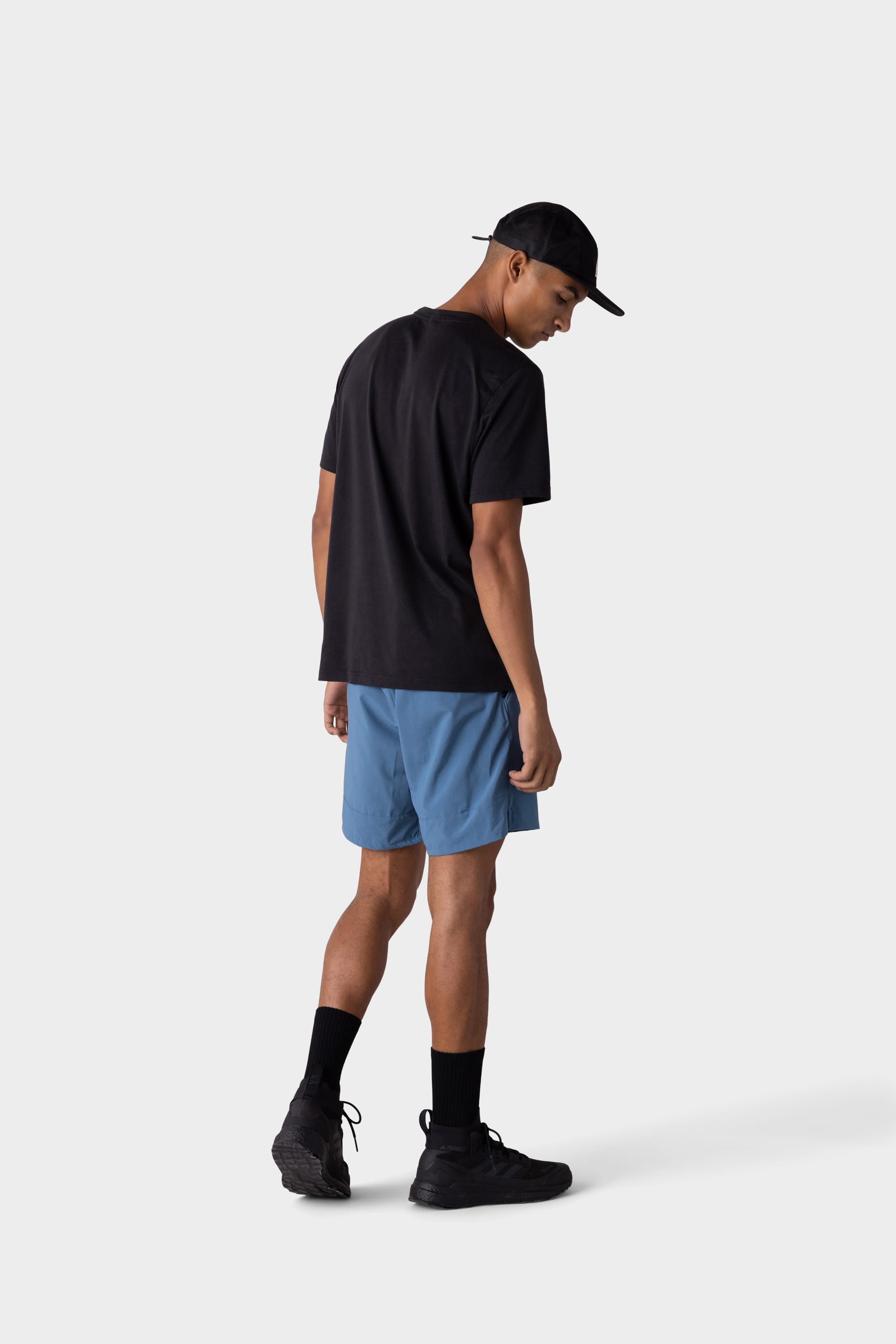 A person in a black cap, black T-shirt, 686 Mens ATP Stretch Performance Short by 686 in blue, black socks, and black sneakers stands facing slightly away against a plain light gray background.
