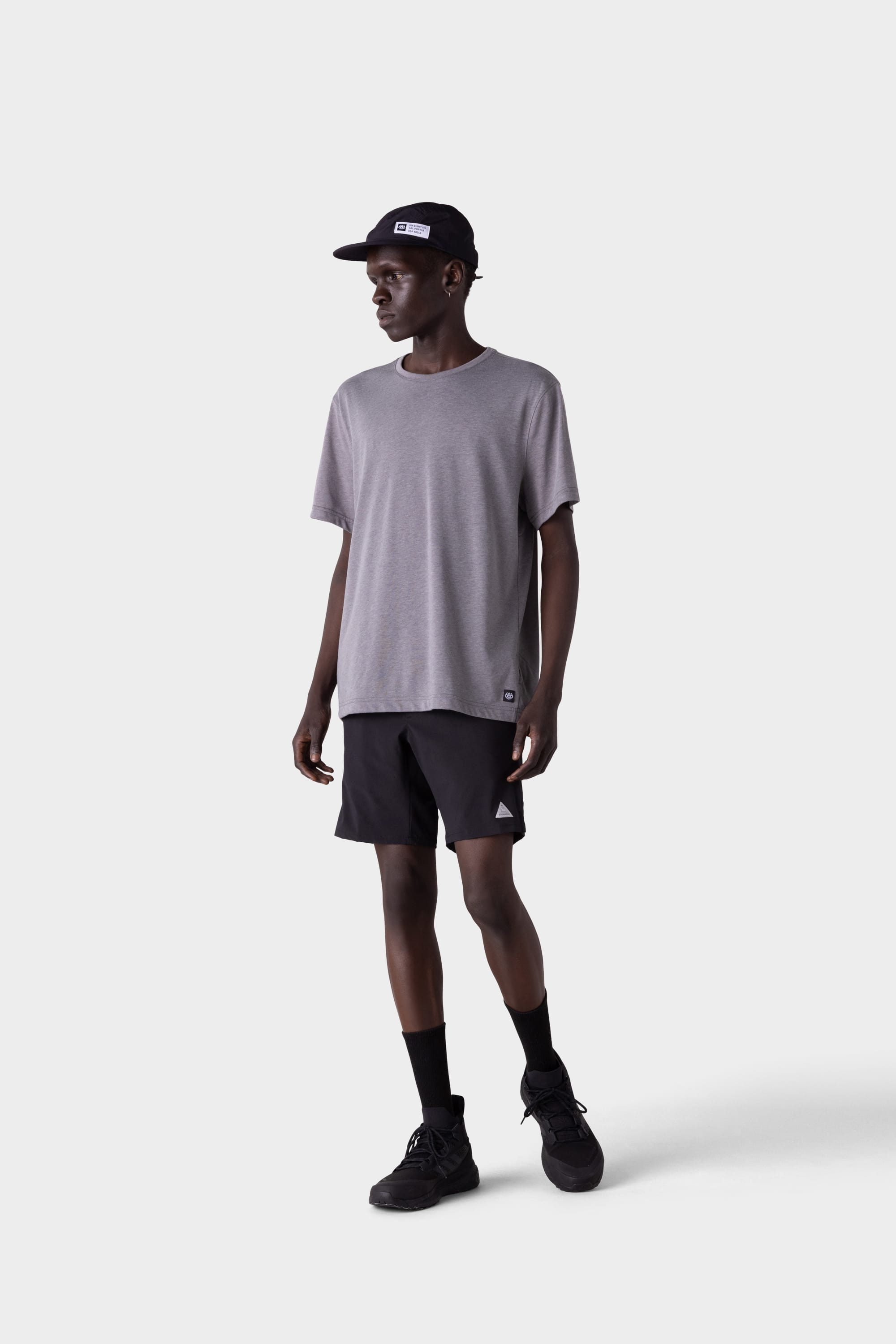 A man wearing a gray t-shirt, black 686 Mens ATP Stretch Performance Short by 686, black sneakers, socks, and cap stands facing slightly to his left against a plain light gray background.
