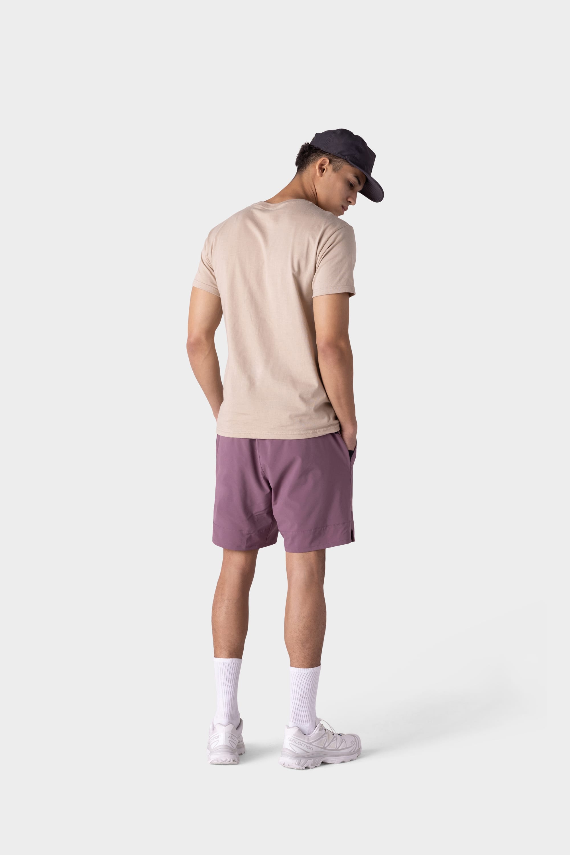A person stands facing away against a plain light background, wearing a beige t-shirt, 686 Mens ATP Stretch Performance Short by 686 in mauve, white crew socks and sneakers, and a dark baseball cap.