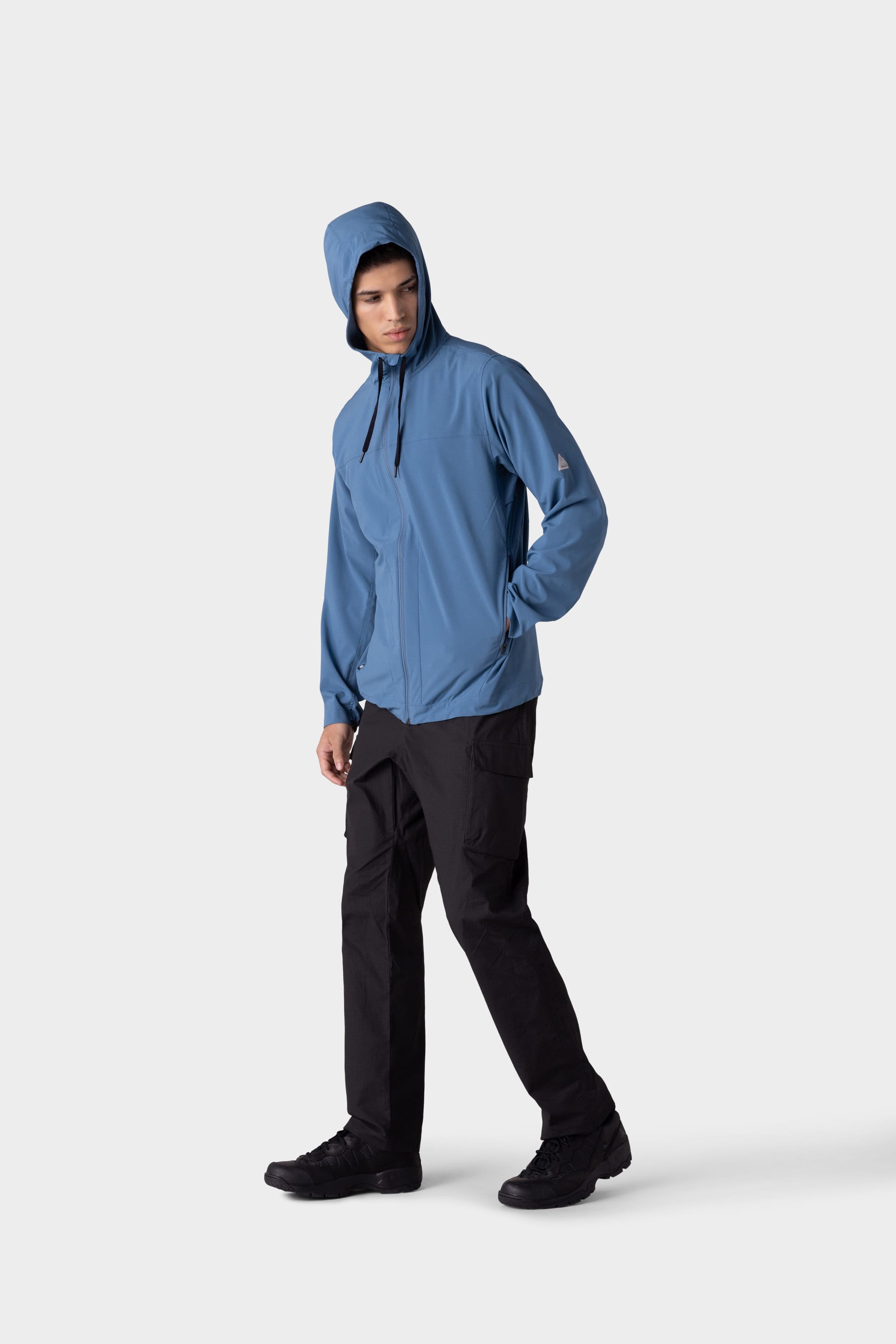 A person wearing the 686 Mens ATP Stretch Wind Jacket in blue, black cargo pants, and black shoes stands against a plain light gray background, looking slightly downward to the left.