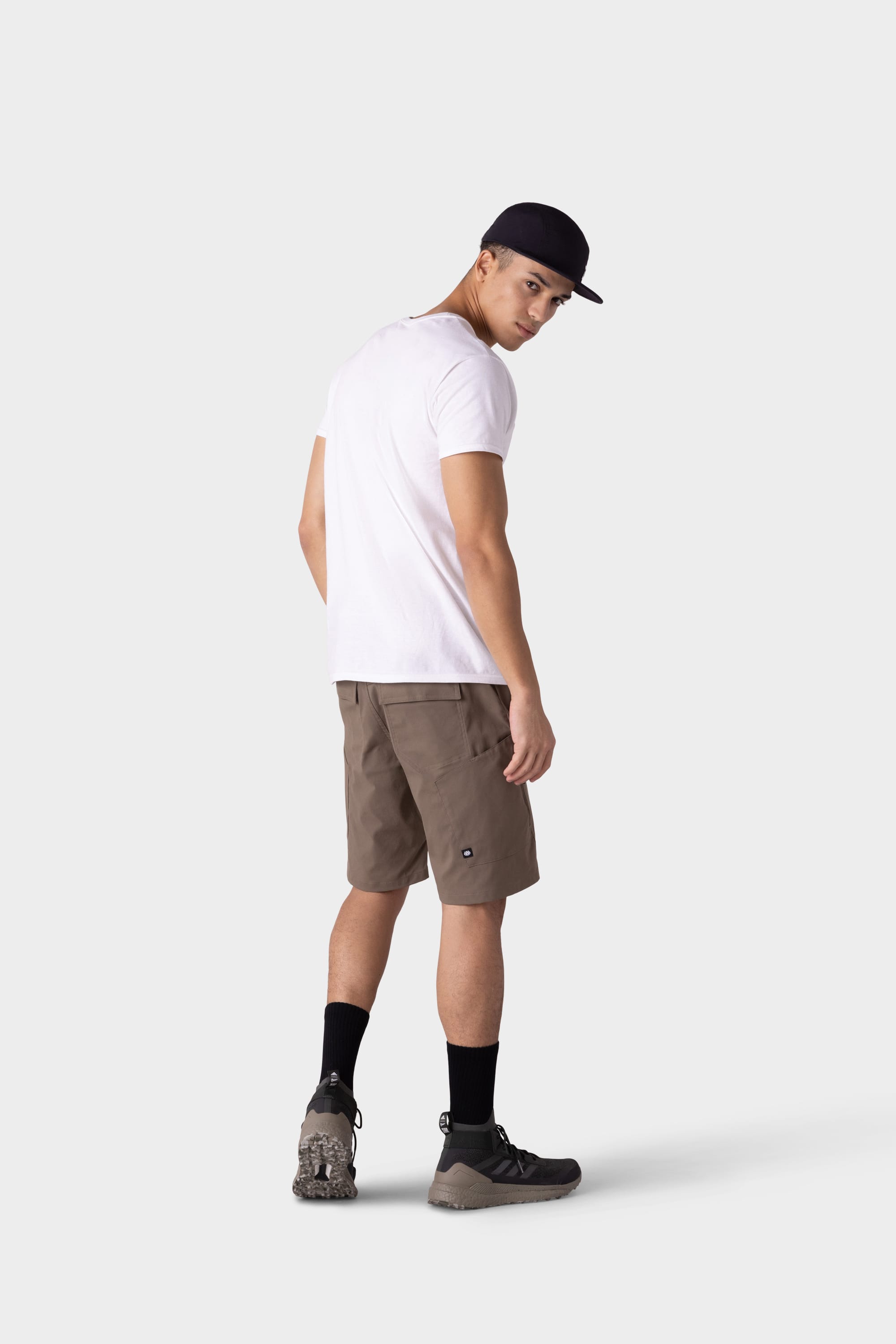 A man models the 686 Mens Anything Hybrid Cargo Short by 686, paired with a white t-shirt, black cap, black socks, and hiking shoes. He stands against a light gray background, looking back to highlight the minimalist style.