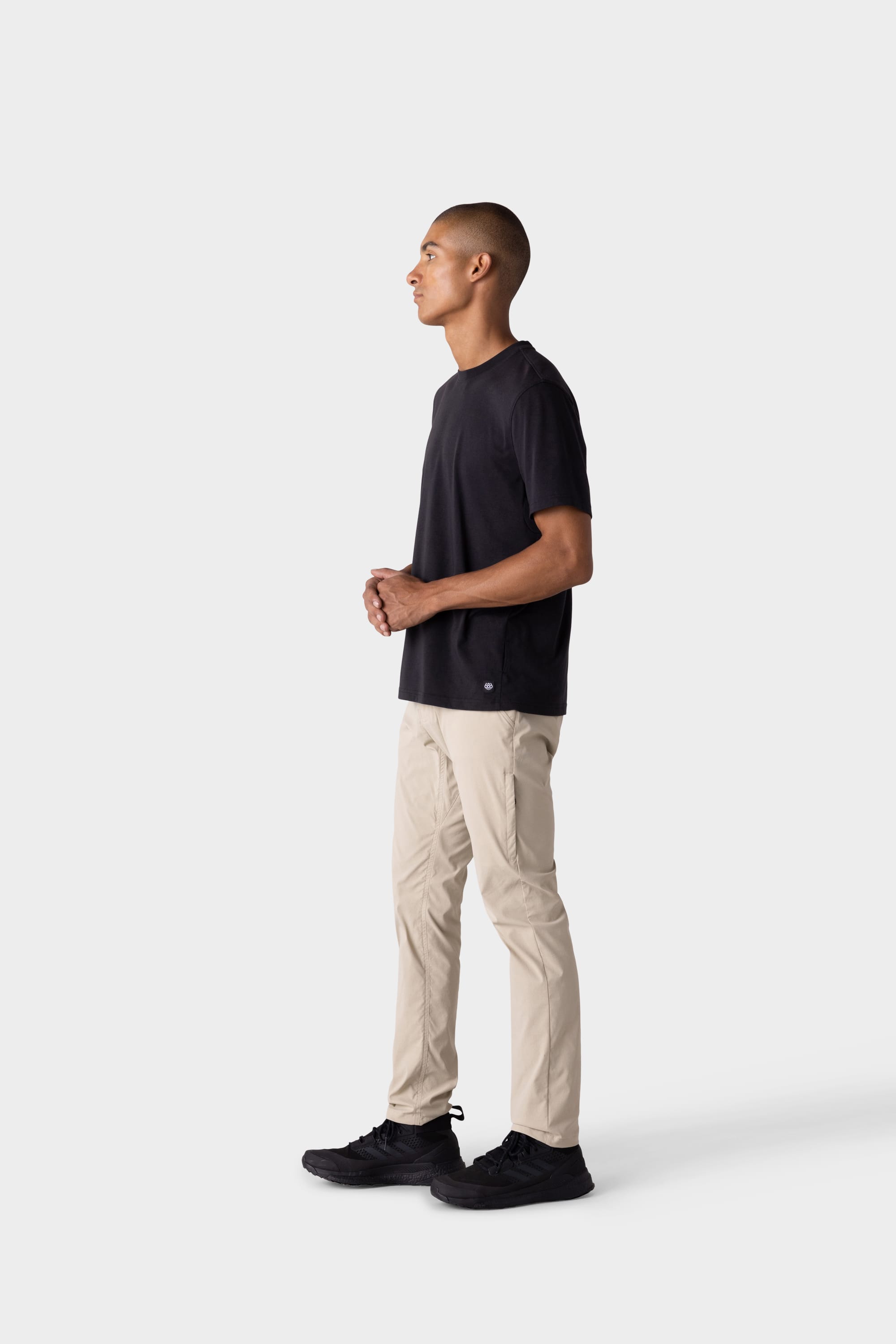 A man stands in profile against a white background, wearing a black t-shirt, black shoes, and 686 Mens Everywhere Featherlight Chino Pant - Slim Fit in light beige. His hands are lightly clasped as he looks ahead, ready for warm weather adventures.