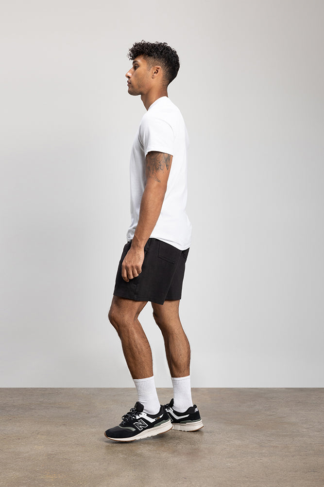 A man with short curly hair stands sideways against a light grey background, wearing a white t-shirt, 686 Mens Everywhere Double Knit Short by 686, white socks, and black sneakers.