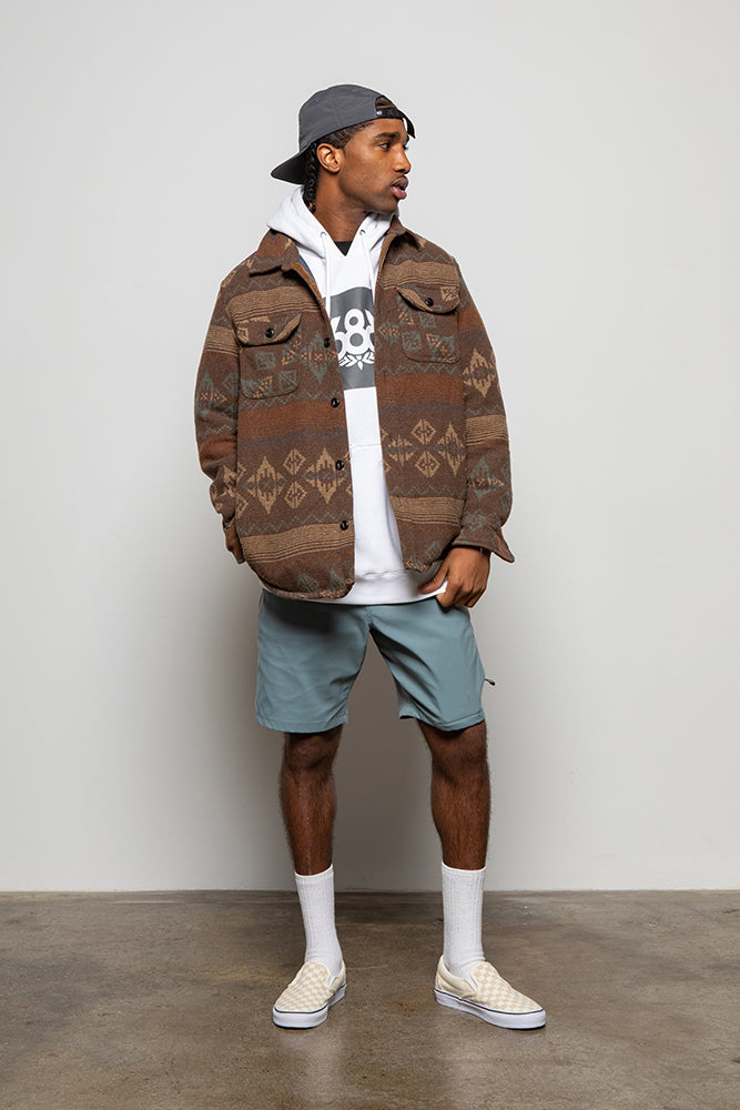 A man stands against a plain wall wearing a gray cap backwards, a brown patterned jacket over a white hoodie, 686 Mens Everywhere Hybrid Short in light blue, white socks, and beige checkered slip-ons. He looks left with one hand in his pocket.