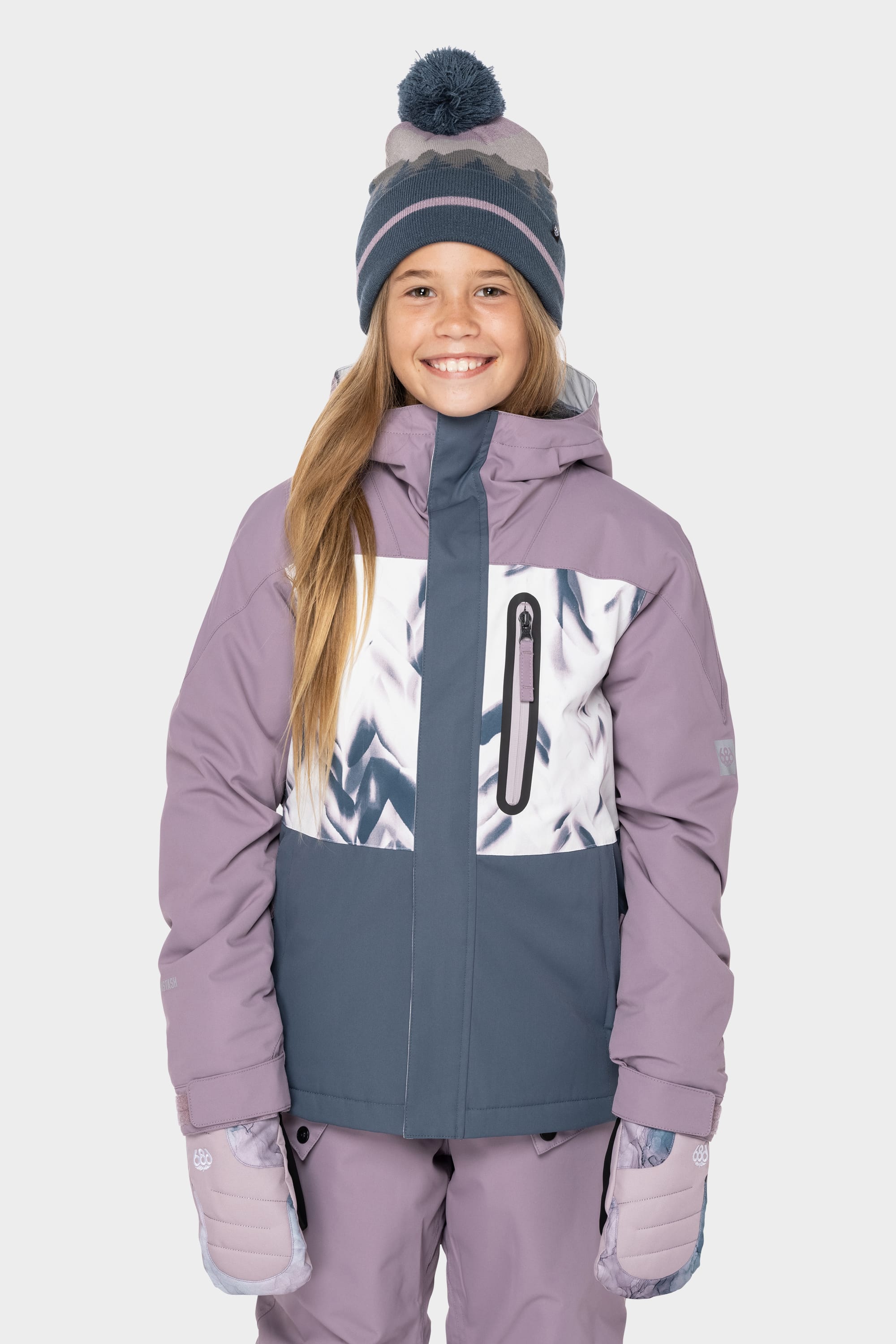 A smiling child wearing the 686 Girls Hydrastash Insulated Jacket by 686, featuring purple and grey colors with a patterned chest panel, matching snow pants, light purple gloves, and a blue pom-pom beanie against a white background.