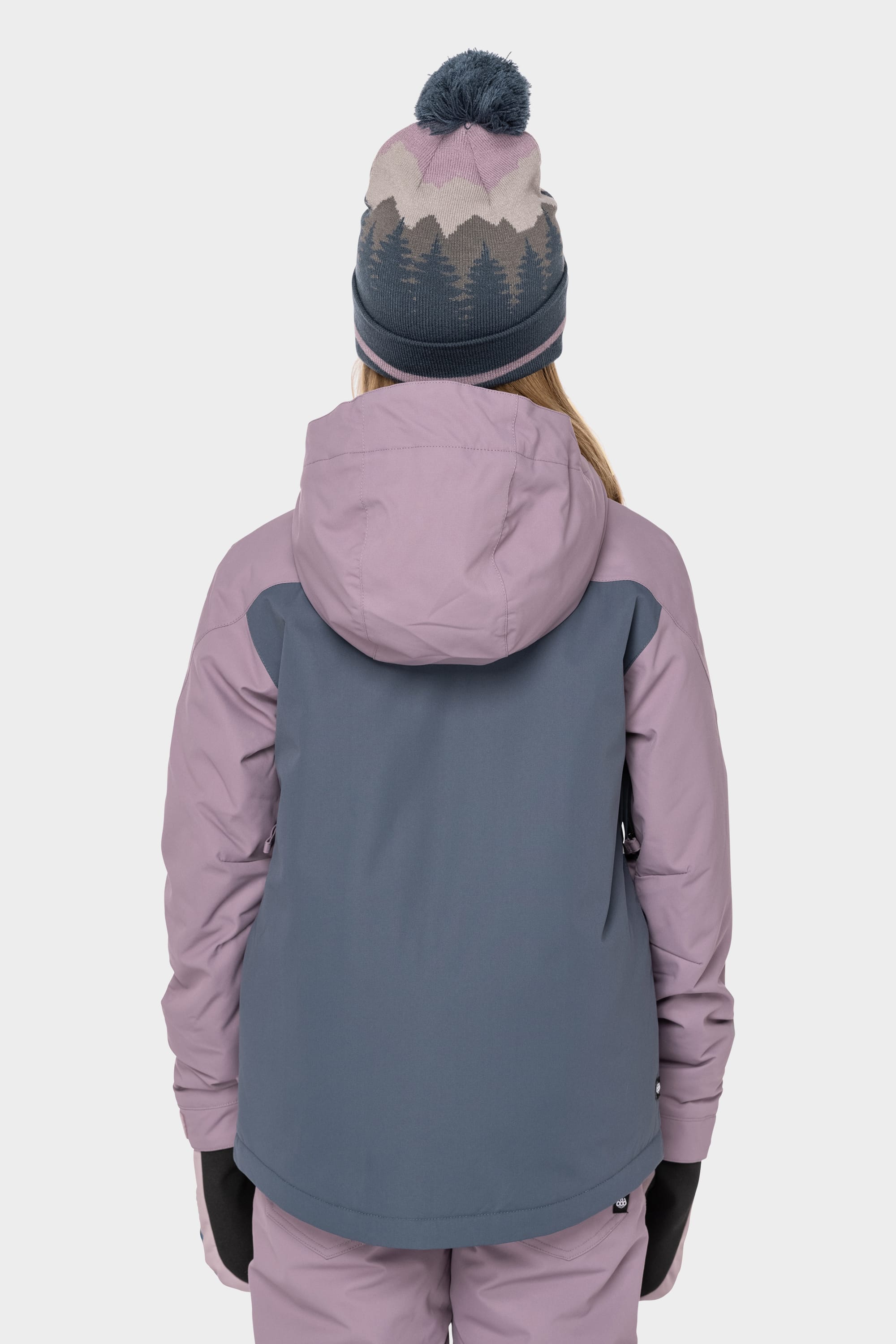 A person is seen from behind wearing the 686 Girls Hydrastash Insulated Jacket in lavender and blue, pink snow pants, black gloves, and a knit beanie with a tree pattern and pom-pom.
