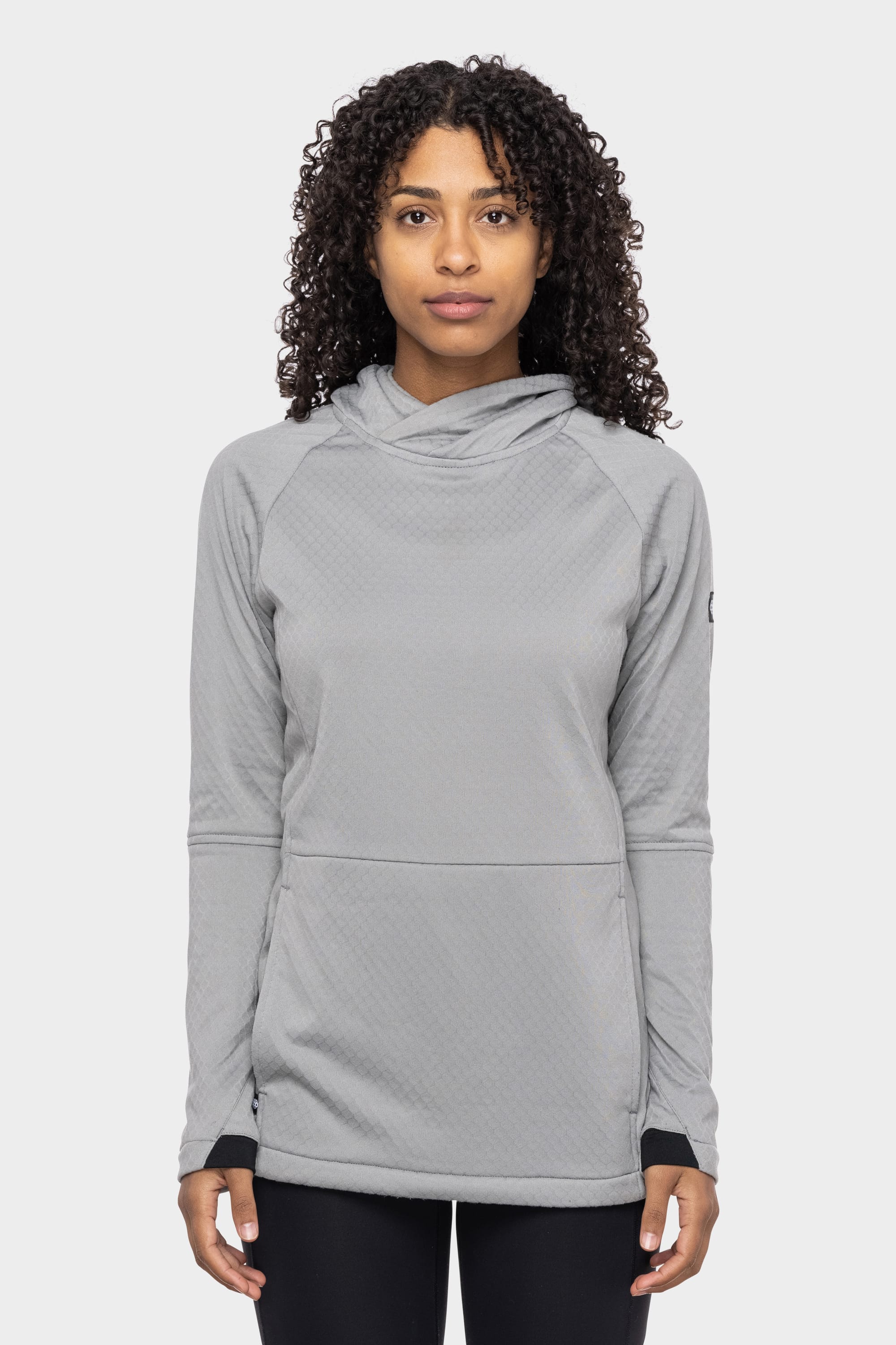 A person with curly hair wears the 686 Womens Mission Grid Fleece Hoody by 686—light grey, long-sleeve with a front pocket—standing against a plain white background.