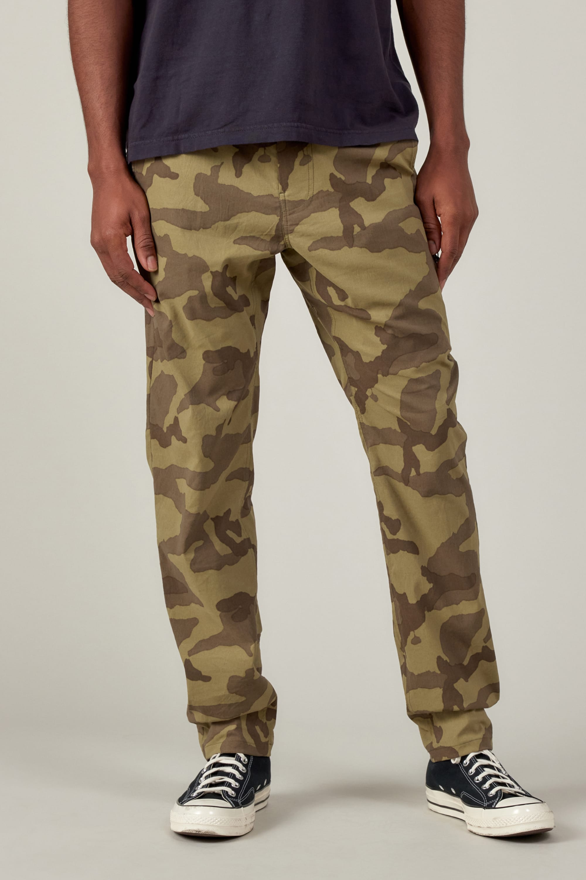 A person in 686 Mens Everywhere Featherlight Chino Pant - Slim Fit, a dark t-shirt, and black low-top sneakers stands against a plain light background—an ideal outfit for warm weather adventures. The image is cropped at the shoulders.