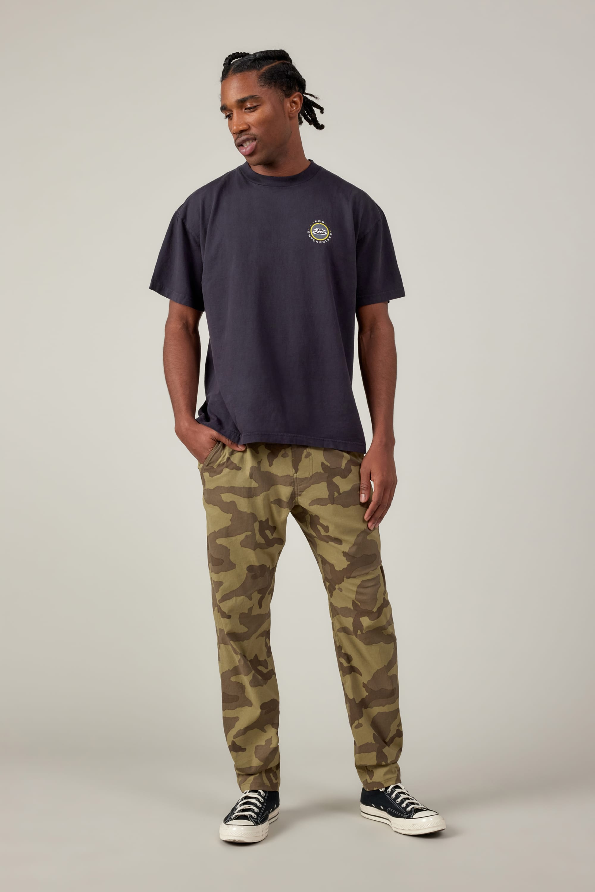 A man stands against a plain background wearing a loose dark tee, 686 Mens Everywhere Featherlight Chino Pant - Slim Fit, and black and white sneakers. His hands are in his pockets as he looks slightly to the side.