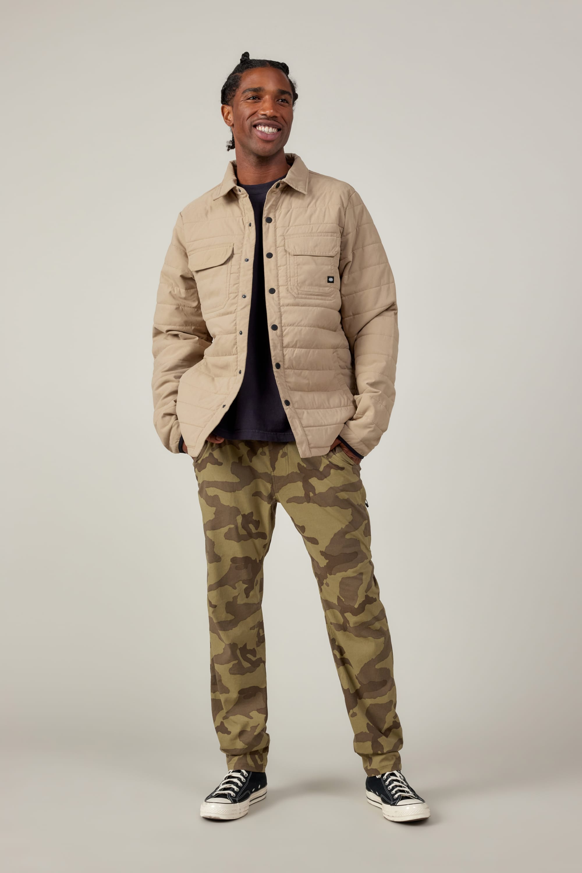 A smiling man stands hands-in-pockets, wearing a beige quilted jacket, black shirt, 686 Mens Everywhere Featherlight Chino Pant - Slim Fit in camouflage print, and black-and-white sneakers—ready for warmer days.