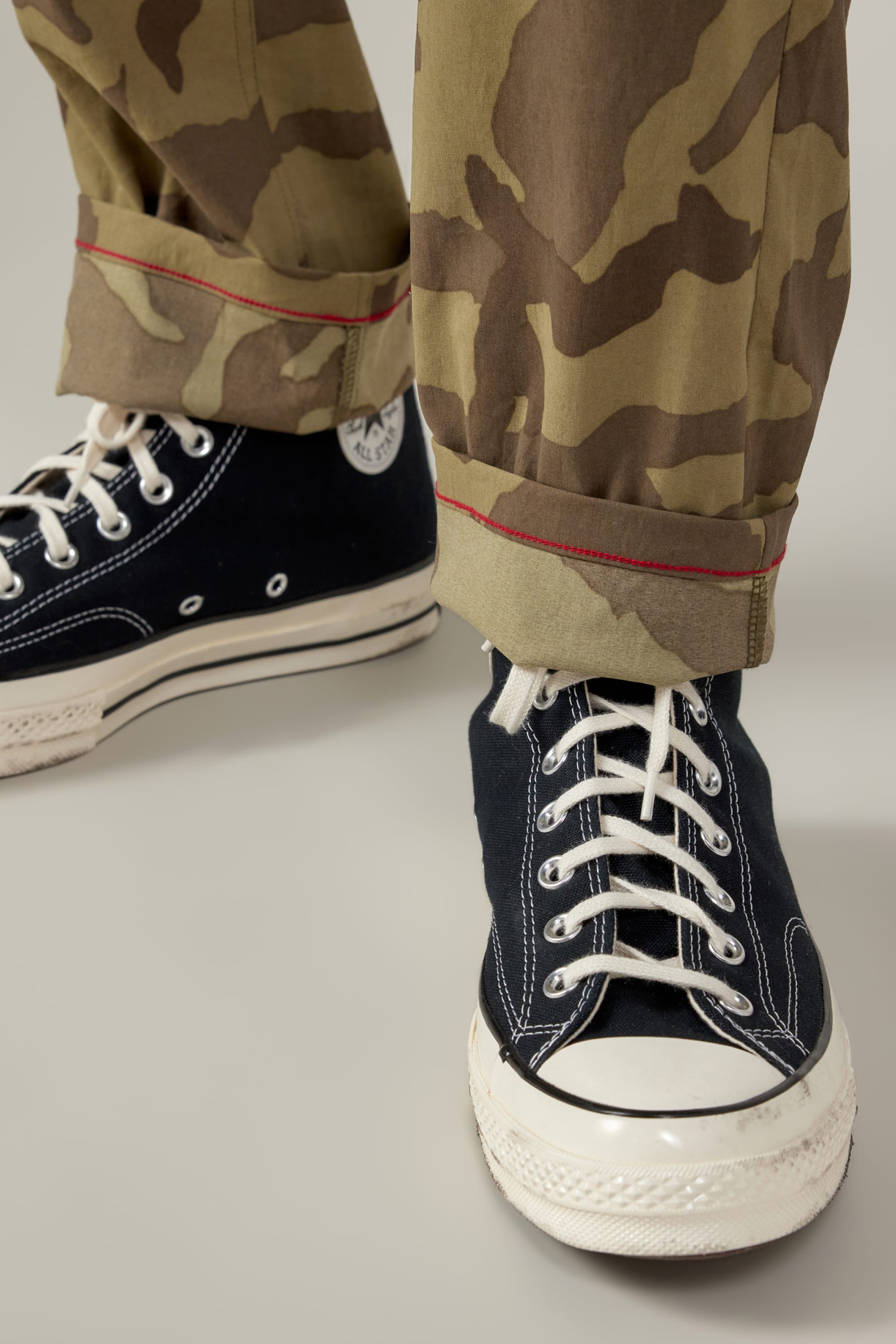 A close-up of someone wearing black high-top Converse and 686 Mens Everywhere Featherlight Chino Pant - Slim Fit, featuring a camo pattern with red stitching and rolled cuffs—ideal for warm-weather adventures, highlighting the pants lightweight fabric.