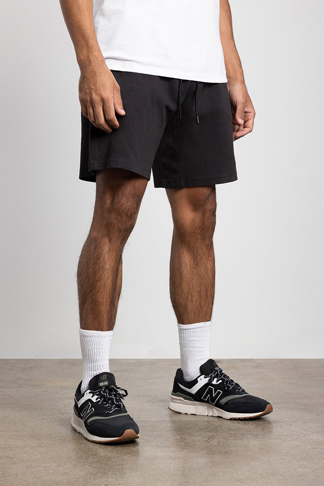 A person wearing a white T-shirt, 686 Mens Everywhere Double Knit Short in black, white crew socks, and black New Balance sneakers stands on a smooth concrete floor against a plain light grey wall.
