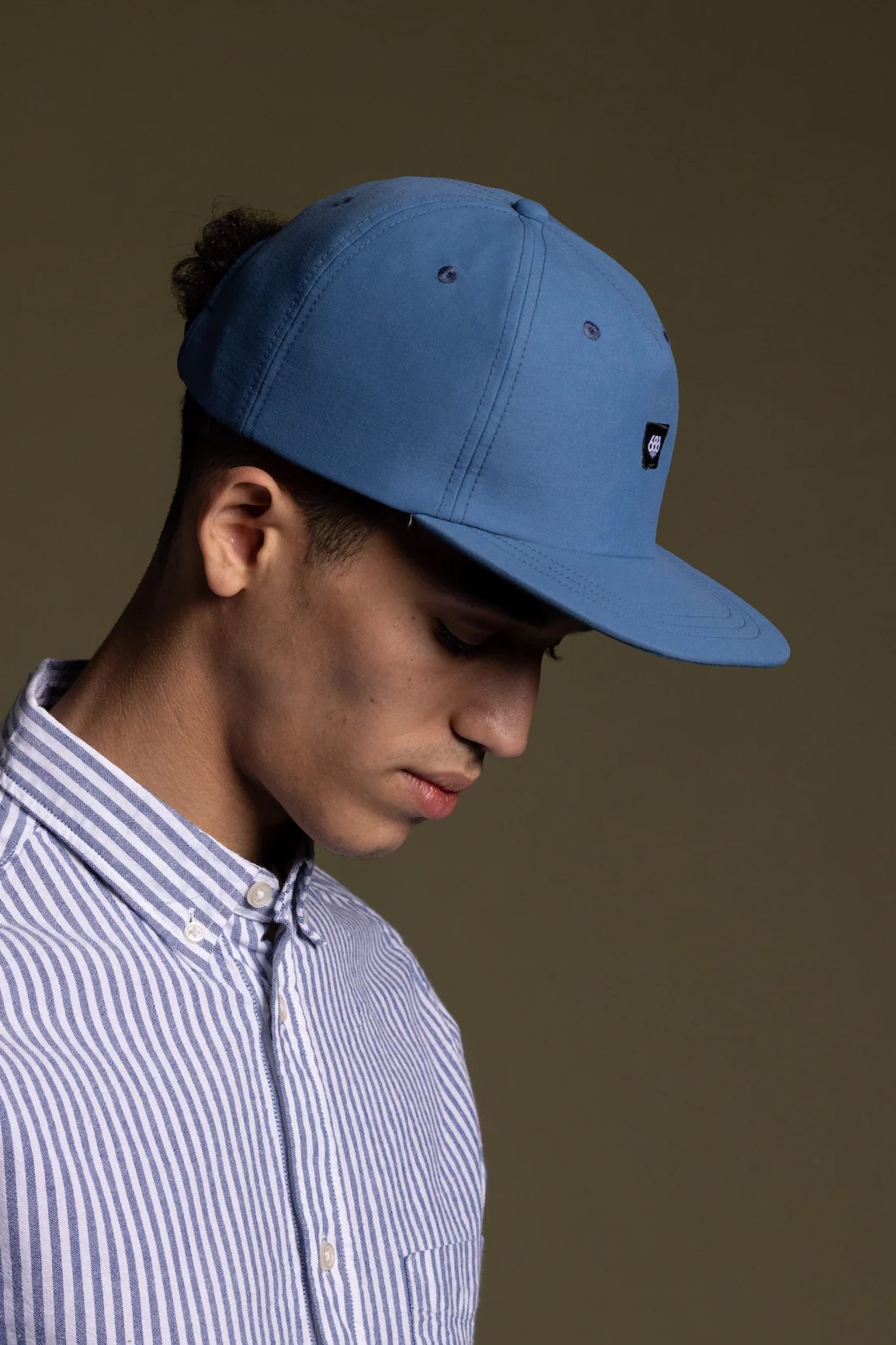 A young person wearing the 686 Packable Everywhere Hat from 686 and a blue-and-white striped button-down shirt looks downward against a plain, neutral background.