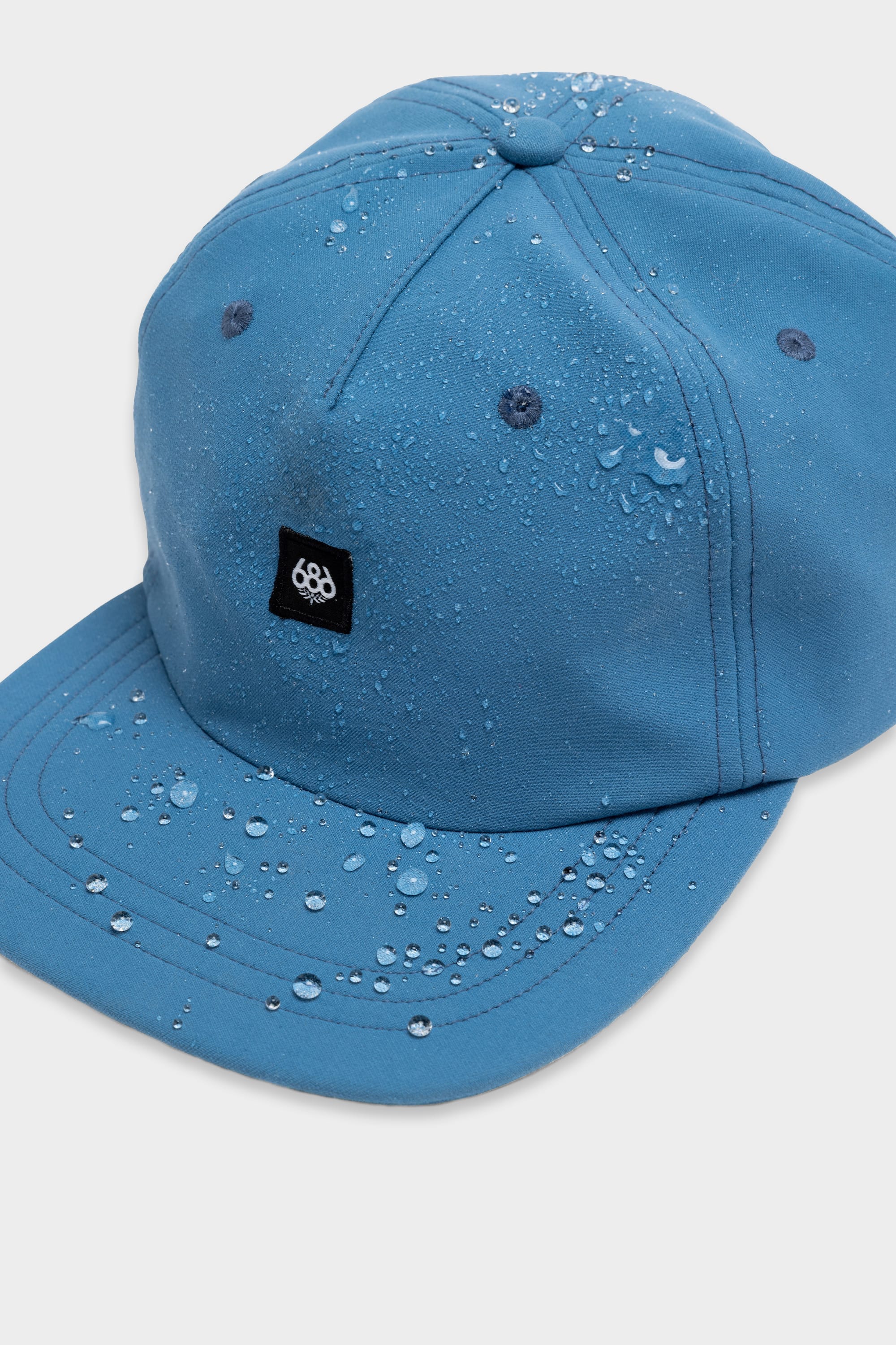 The 686 Packable Everywhere Hat by 686 is a blue, water-resistant baseball cap with small droplets on its surface. It features a small black patch with a white logo on the front and is shown against a plain white background.