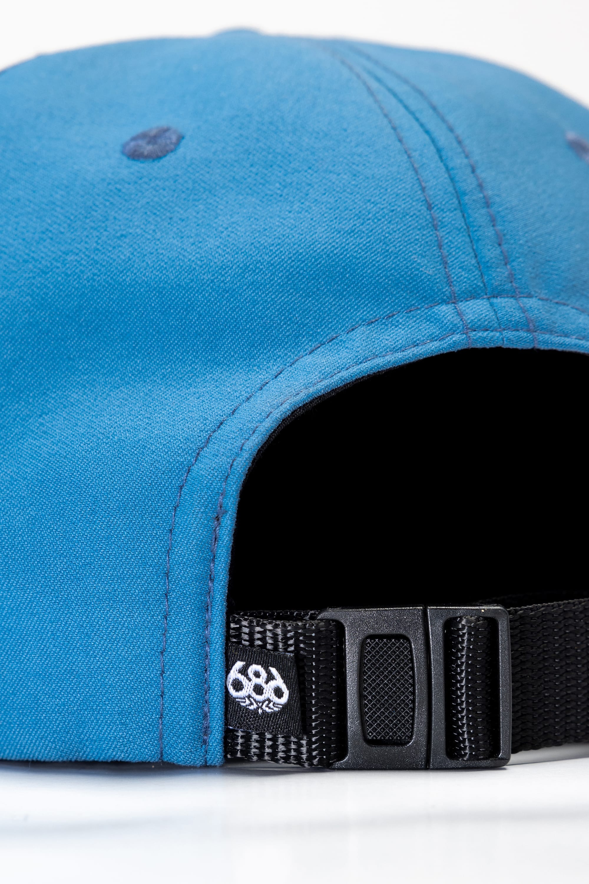 Close-up of the back of the 686 Packable Everywhere Hat in blue, featuring a black adjustable strap with a plastic buckle and a small white embroidered 686 logo near the strap.