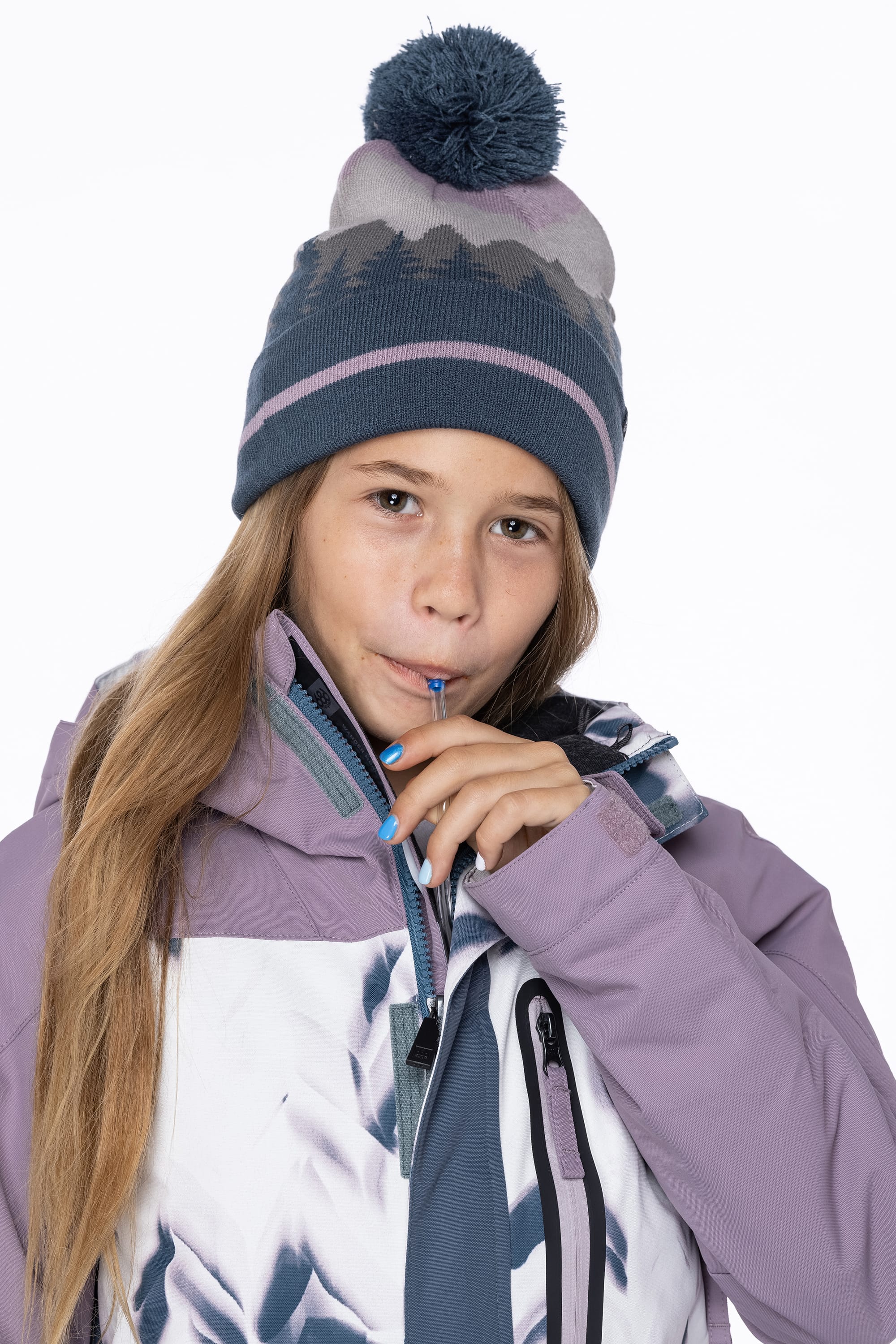 A young girl wearing the 686 Girls Hydrastash Insulated Jacket sips from a hydration tube, facing the camera in a blue knit hat with a pom-pom against a plain white background.