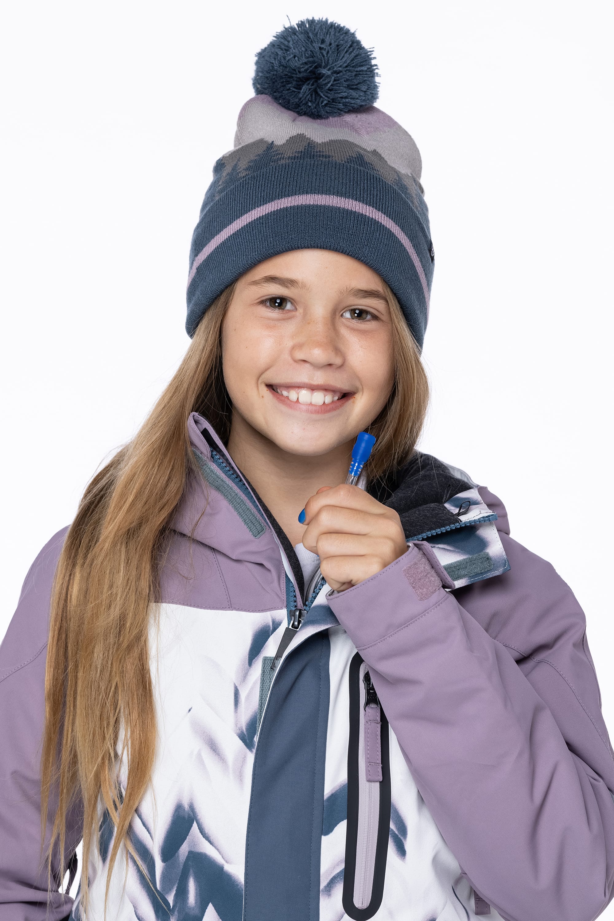 A smiling young girl with long blonde hair wears the 686 Girls Hydrastash Insulated Jacket and a knit beanie with a pom-pom, holding a blue pen near her chin against a white background.