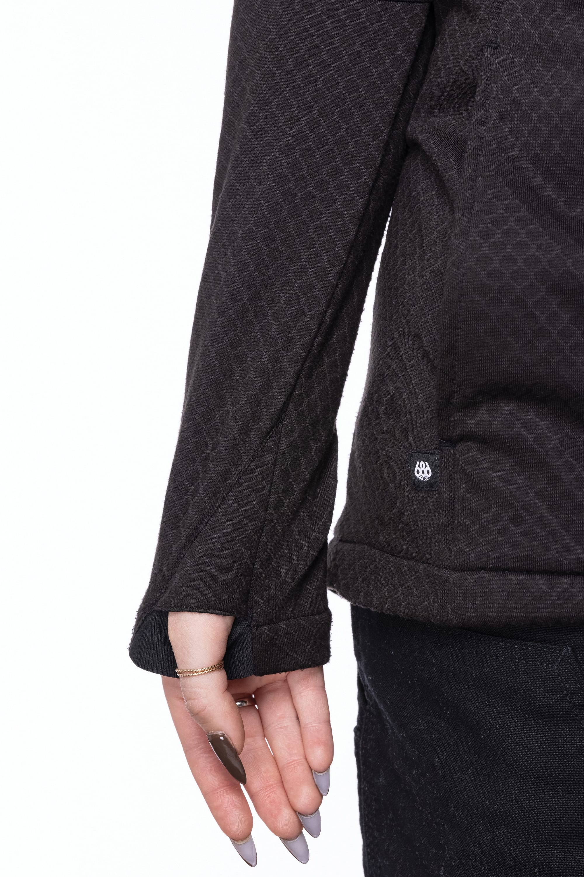 A person wearing the 686 Womens Mission Grid Fleece Hoody with a thumbhole detail and black pants, showing just the lower arm, hand with long painted nails, and part of the side against a white background.