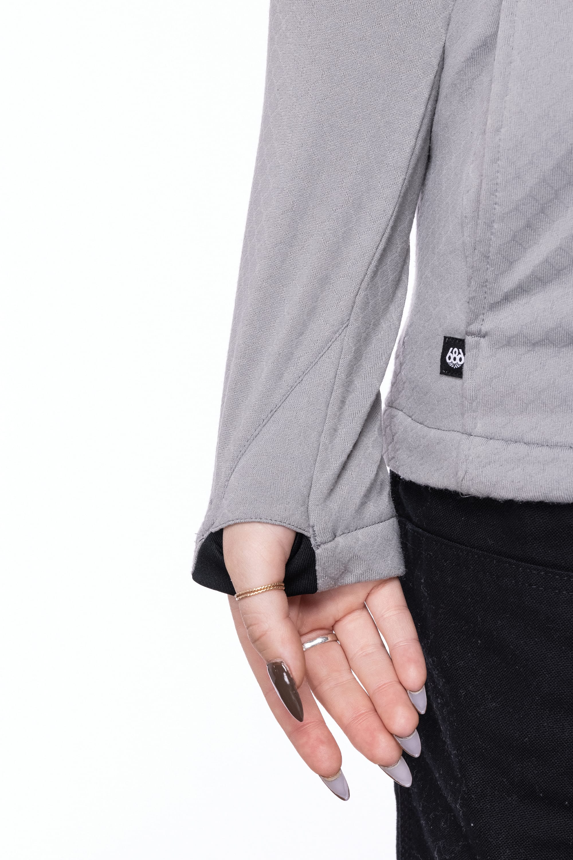 A person with long, manicured nails and a delicate ring holds the hem of a light grey 686 Womens Mission Grid Fleece Hoody against a white background, showing only part of the torso and arm.