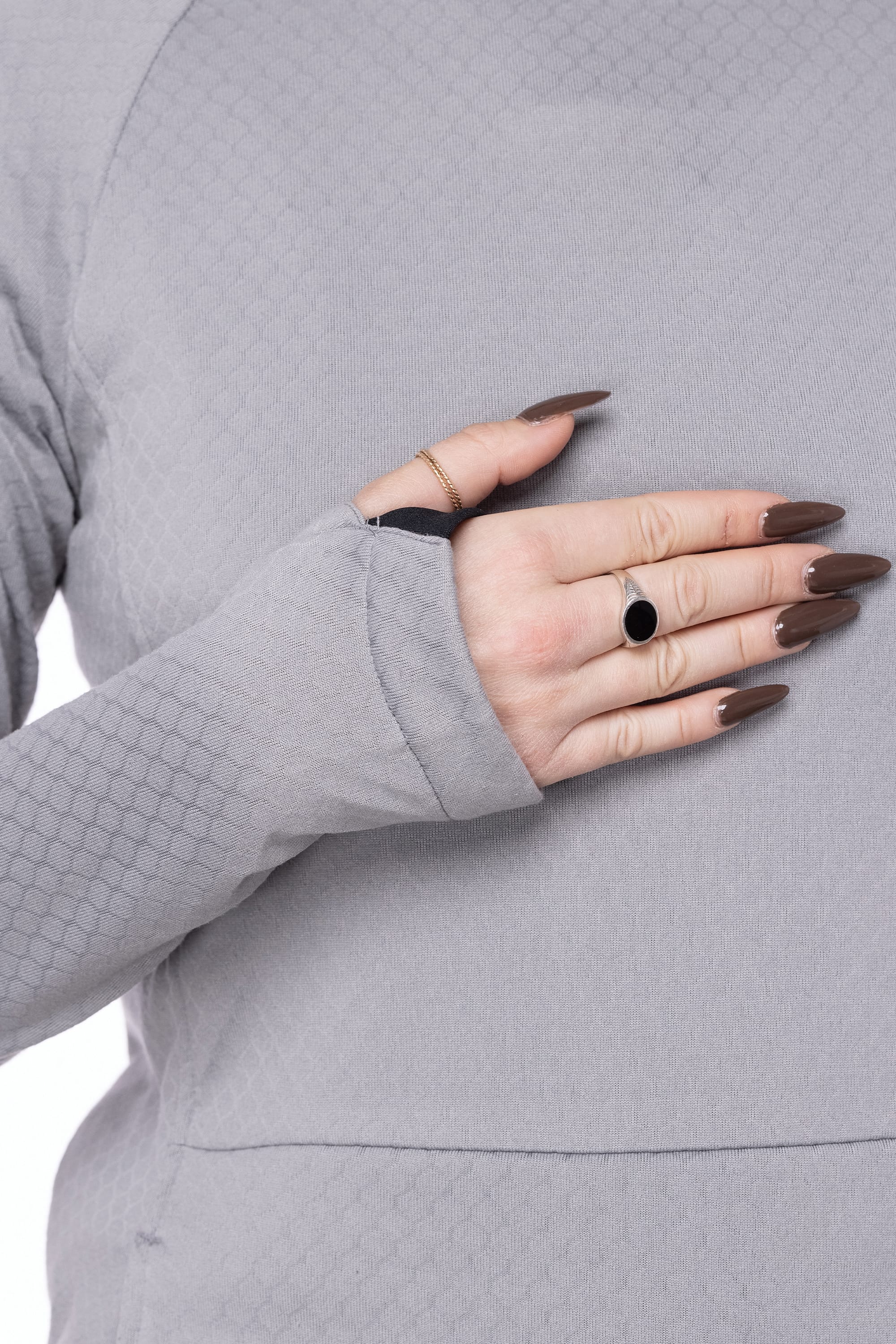 A close-up shows a person wearing the 686 Womens Mission Grid Fleece Hoody in light grey, featuring textured long sleeves with thumbholes. Their hand, with brown manicured nails and rings, gently rests on their chest.