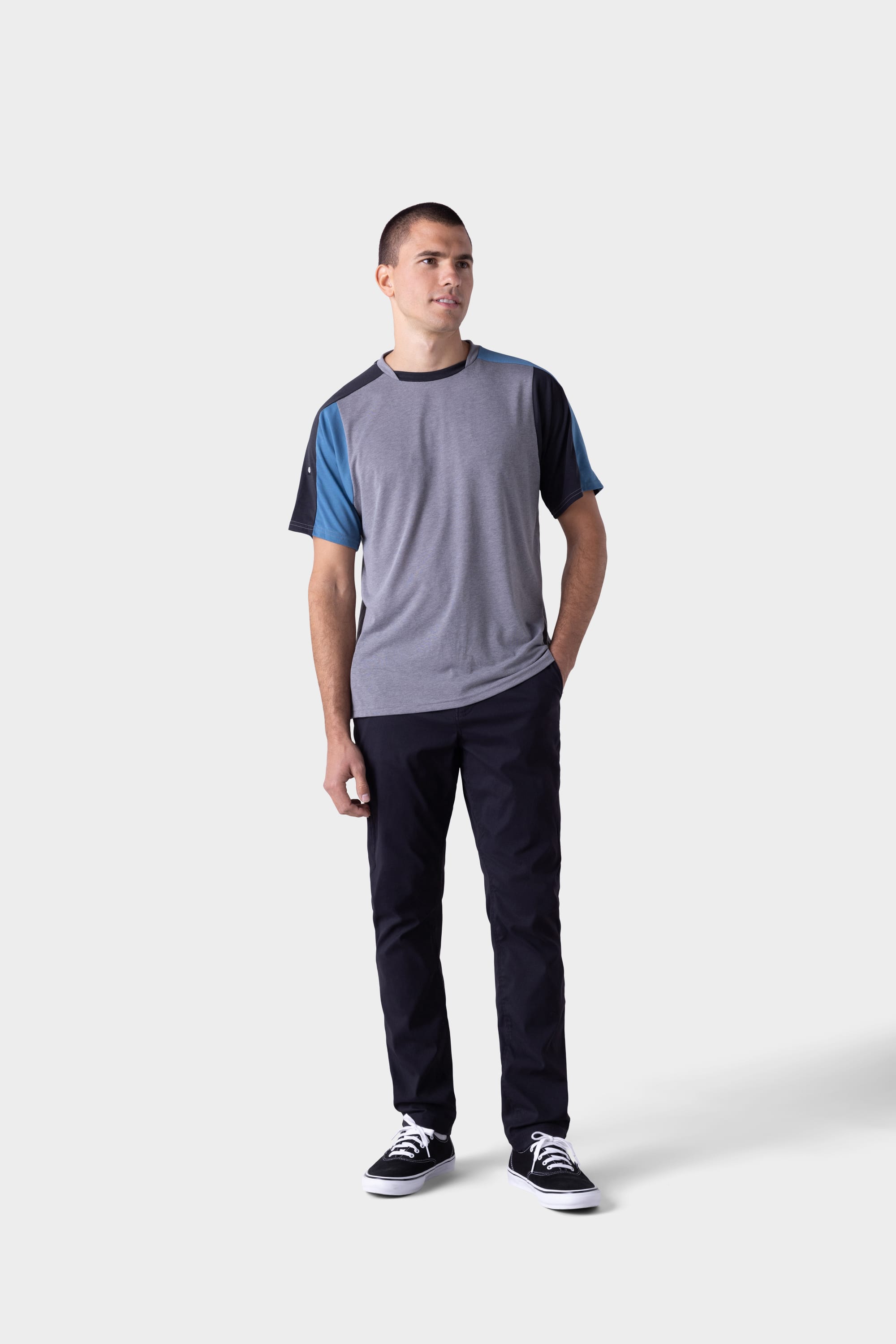 A man stands against a light grey background, wearing a color-blocked grey and blue tee, black sneakers with white soles, and 686 Mens Platform Bike Pant - Slim Fit by 686. One hand is in his pocket as he looks slightly to the side.