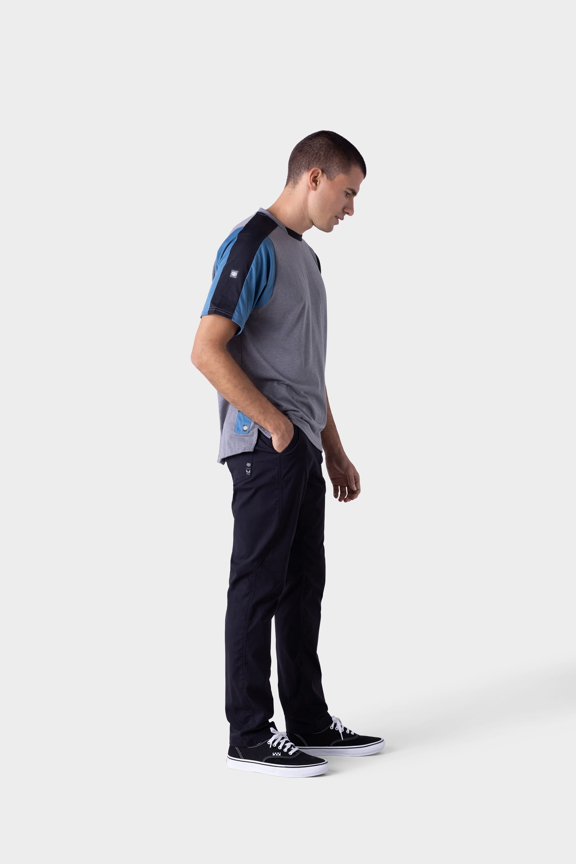 A young man stands against a plain light background, wearing a grey and blue short-sleeve t-shirt, black 686 Mens Platform Bike Pant - Slim Fit, and black sneakers with white soles. He looks down with his hands in his pockets.
