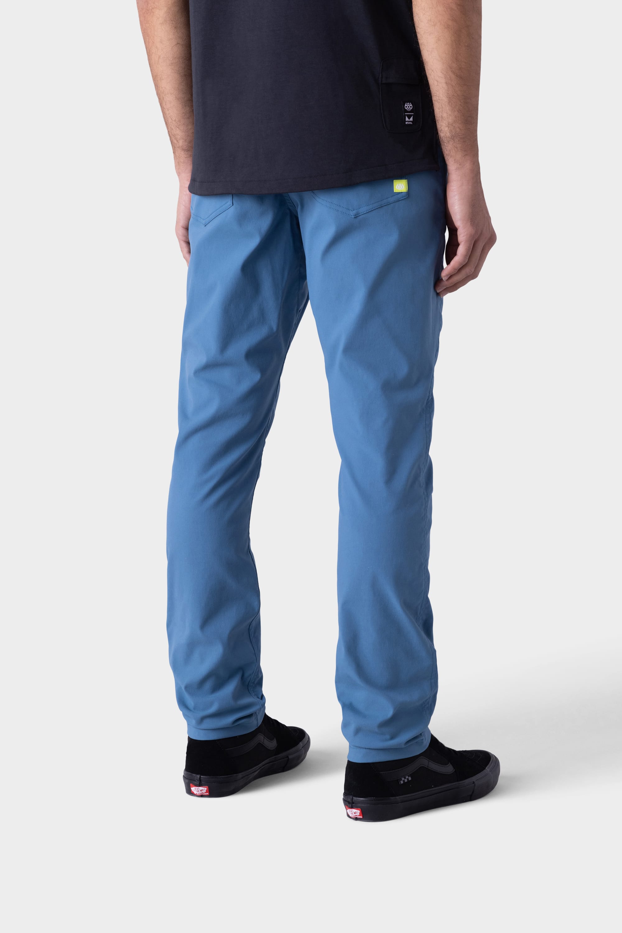 A person stands facing away, wearing 686 Mens Platform Bike Pant - Slim Fit in light blue, a black t-shirt, and black sneakers with white soles. The background is plain and light-colored.