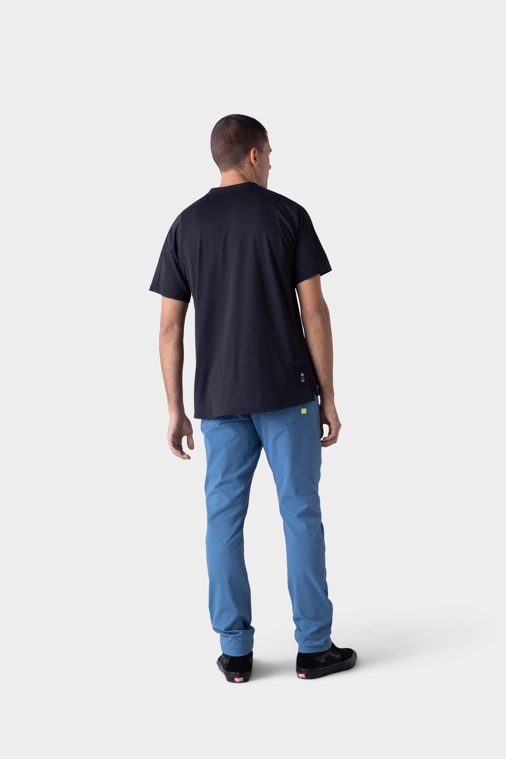 A man faces away against a light grey background, wearing a black T-shirt, black sneakers, and 686 Mens Platform Bike Pant - Slim Fit by 686 in blue.