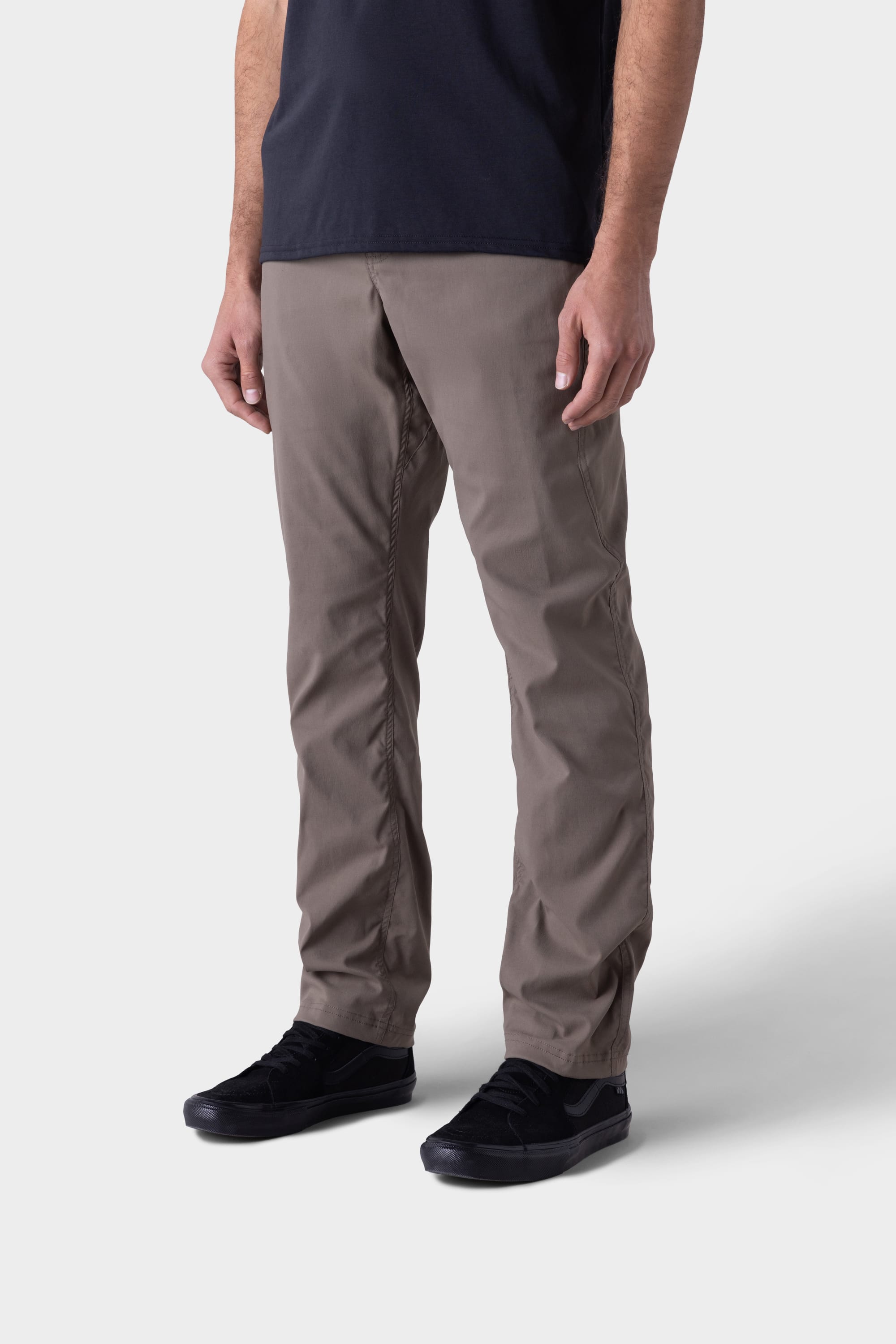 A man wearing the 686 Mens Platform Bike Pant - Relaxed Fit in taupe stands against a plain light grey background, shown from shoulders to feet, paired with a black T-shirt and black sneakers.