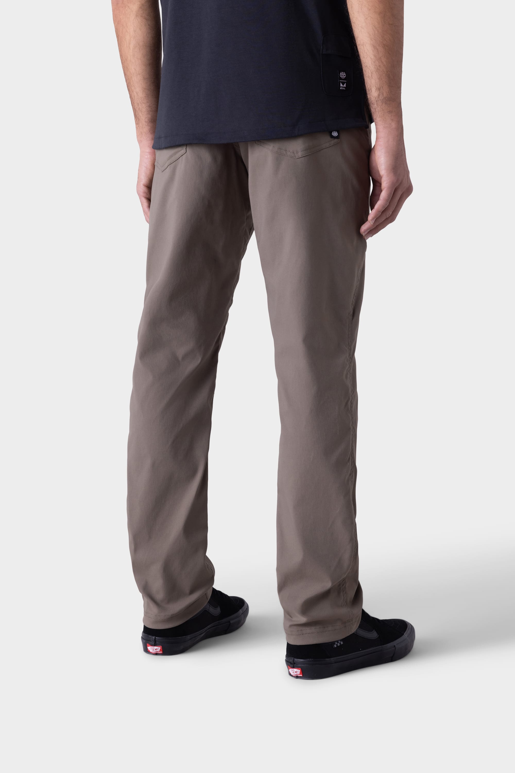 A person stands facing away, wearing 686 Mens Platform Bike Pant - Relaxed Fit in taupe, a black t-shirt, and black sneakers with white soles, against a plain light grey background.