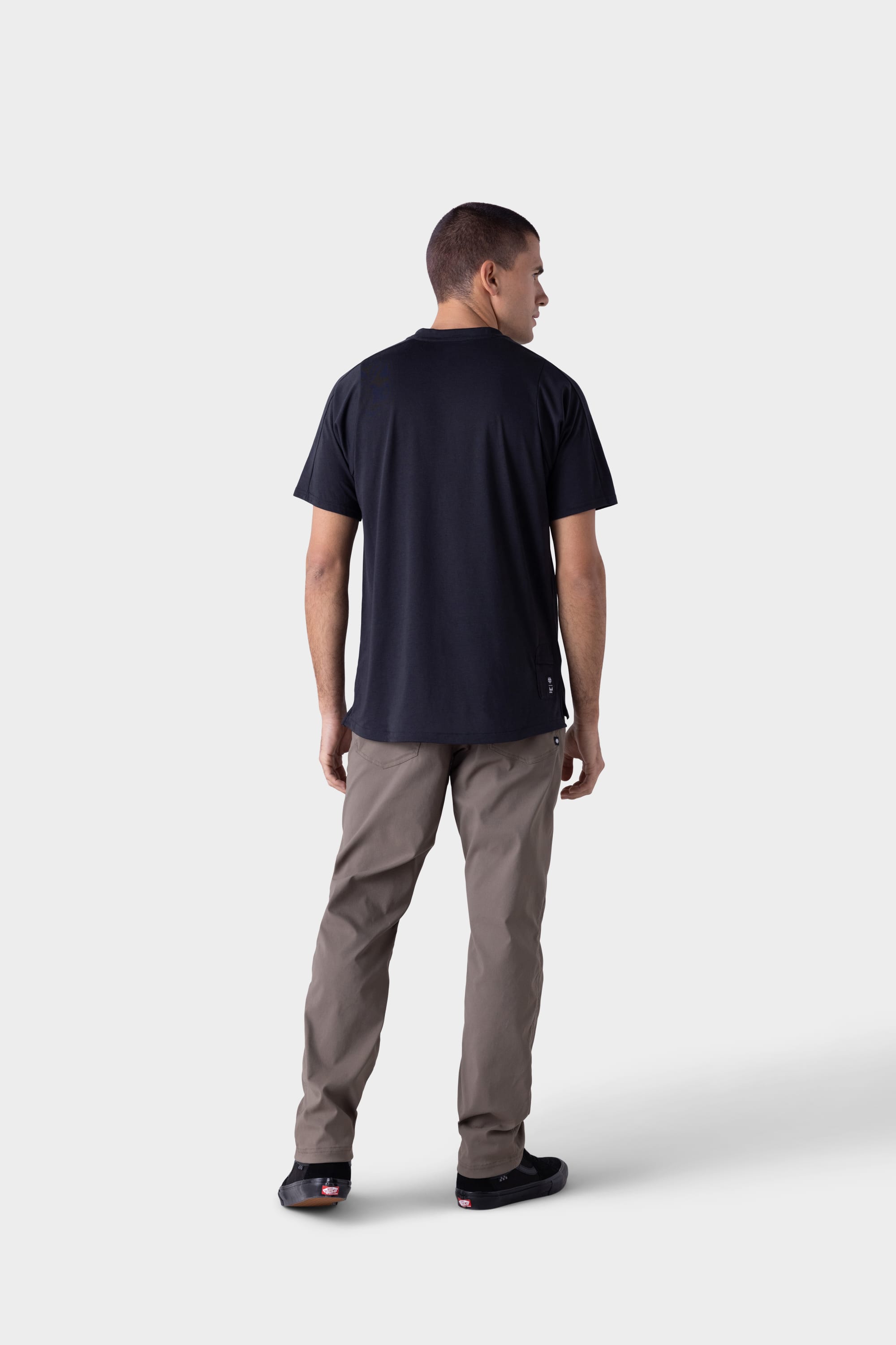 A man faces away from the camera, wearing 686 Mens Platform Bike Pant - Relaxed Fit in light brown, a black t-shirt, and black shoes against a plain light grey background.
