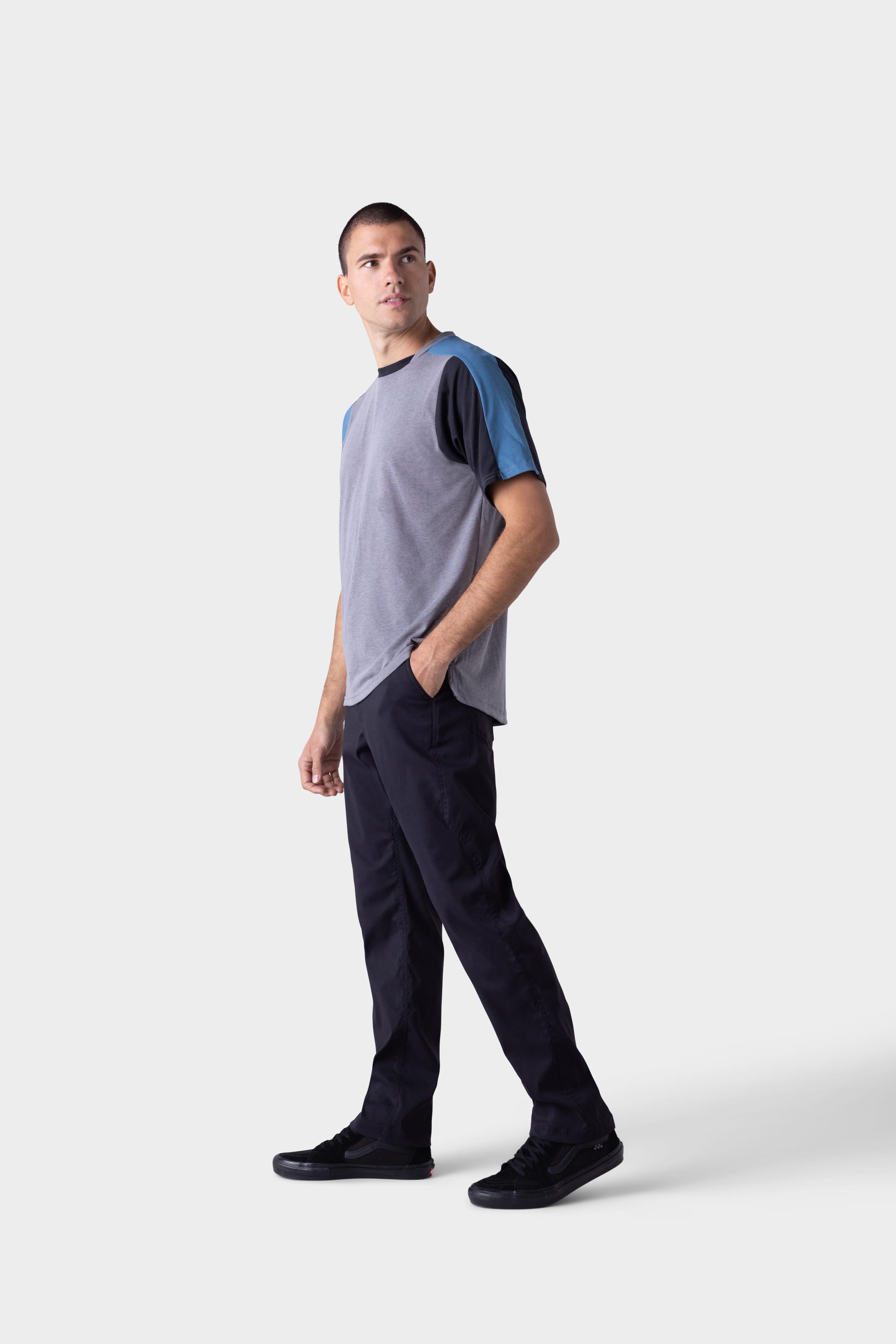 A man stands against a plain light grey background, wearing black 686 Mens Platform Bike Pant - Relaxed Fit, a short-sleeve color-block t-shirt with grey and blue sleeves, black shoes, and looking off to his left.