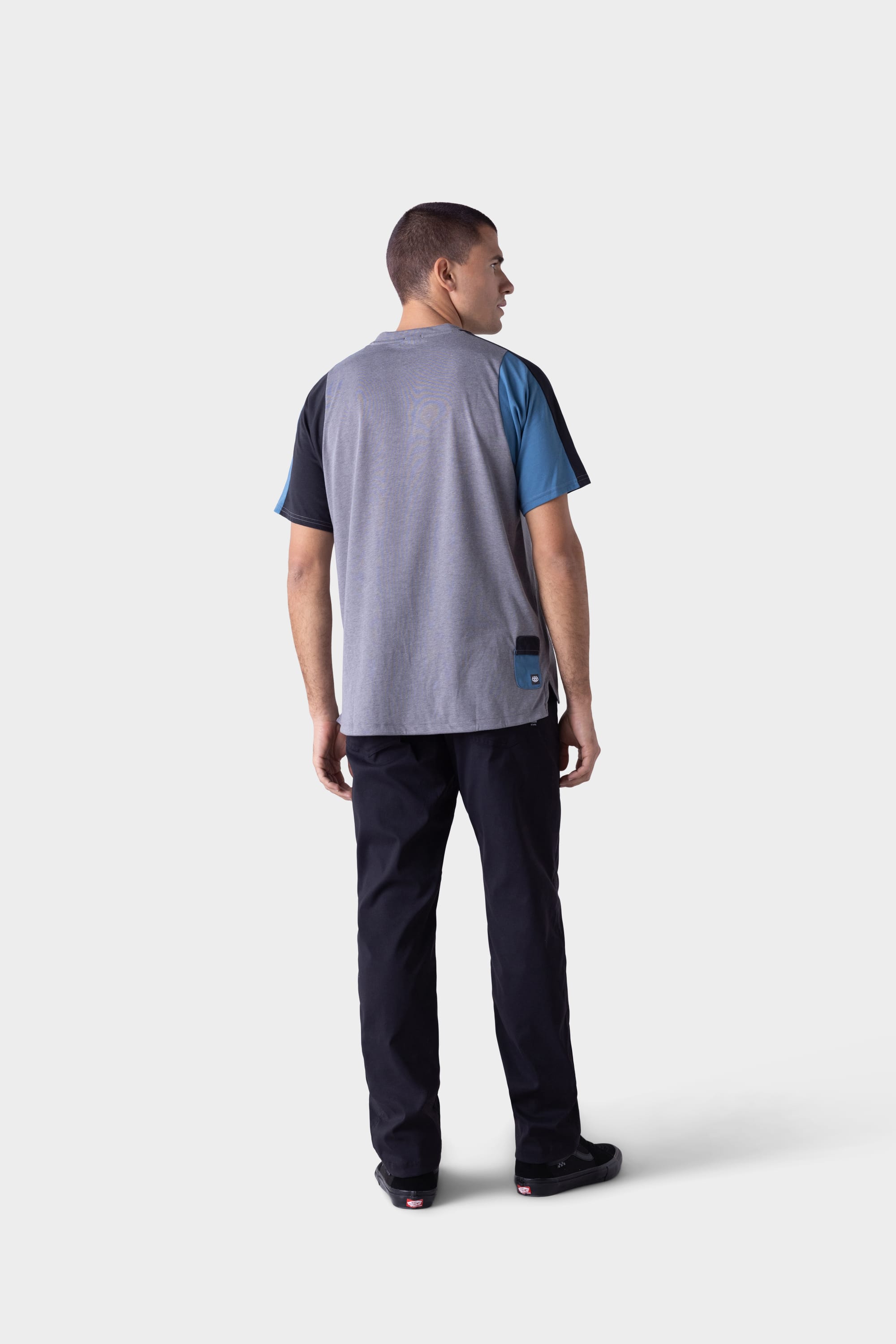 A man faces away in a loose grey tee with blue and black sleeves, paired with black 686 Mens Platform Bike Pant - Relaxed Fit by 686, and black shoes, set against a plain light background.