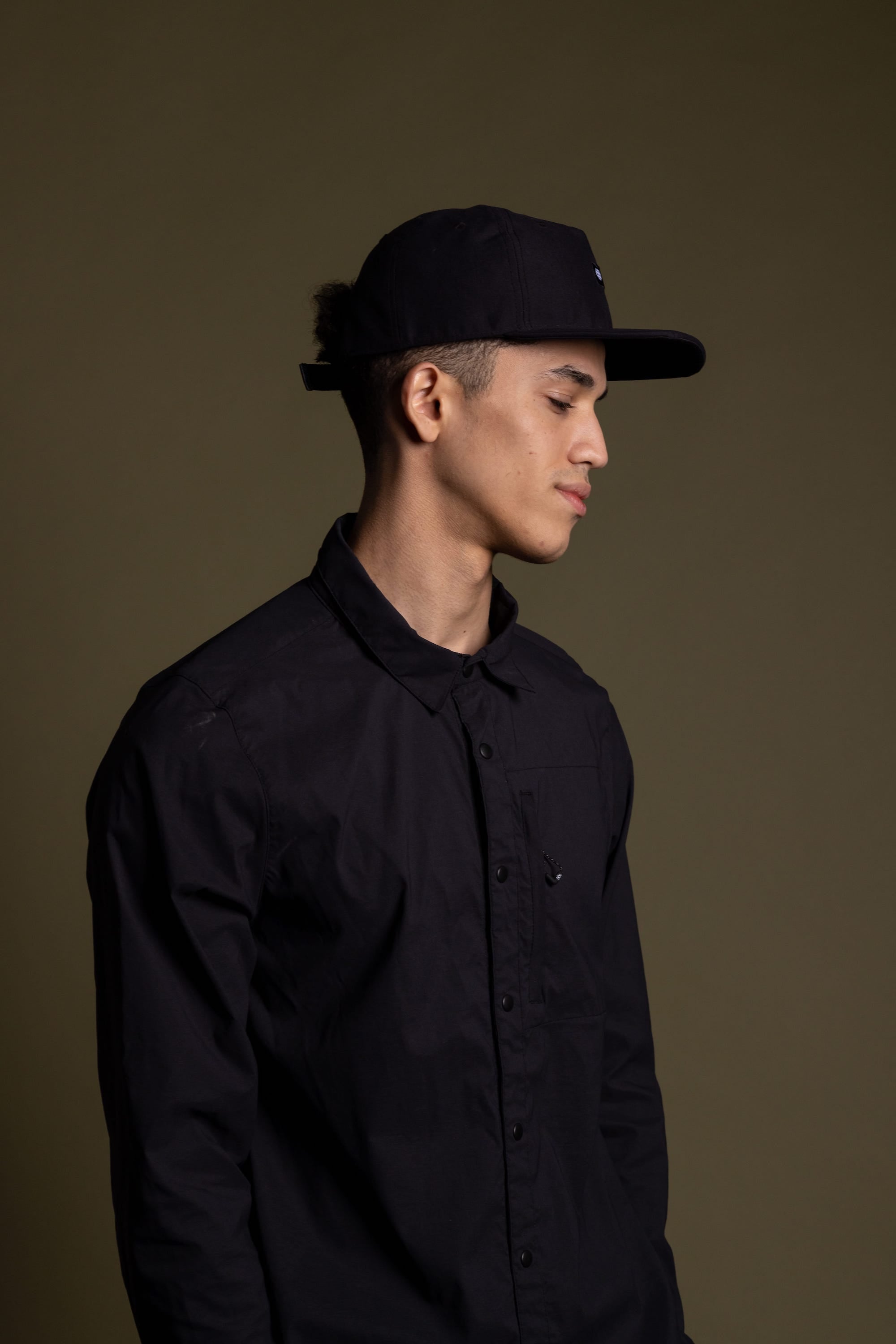 A young man wearing a black button-up shirt and the 686 Packable Everywhere Hat by 686 stands against a plain olive green background, facing right with a neutral expression.